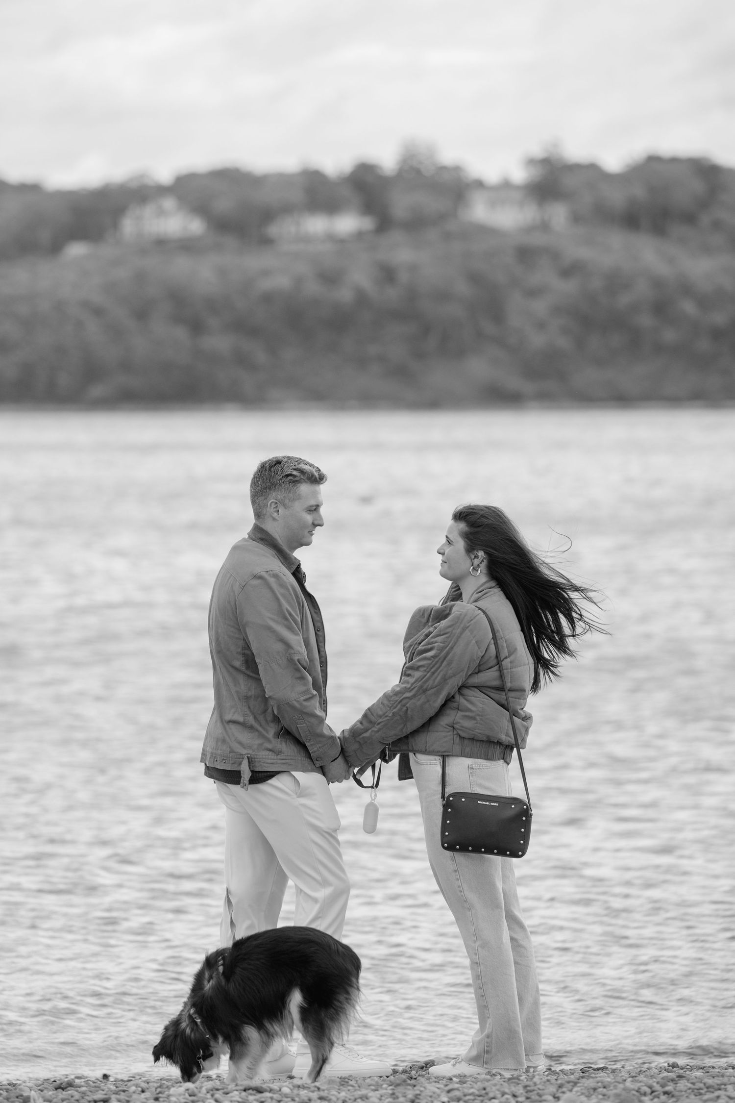 A couple and their dog enjoy a windy day by the water's edge in a romantic black and white scene.