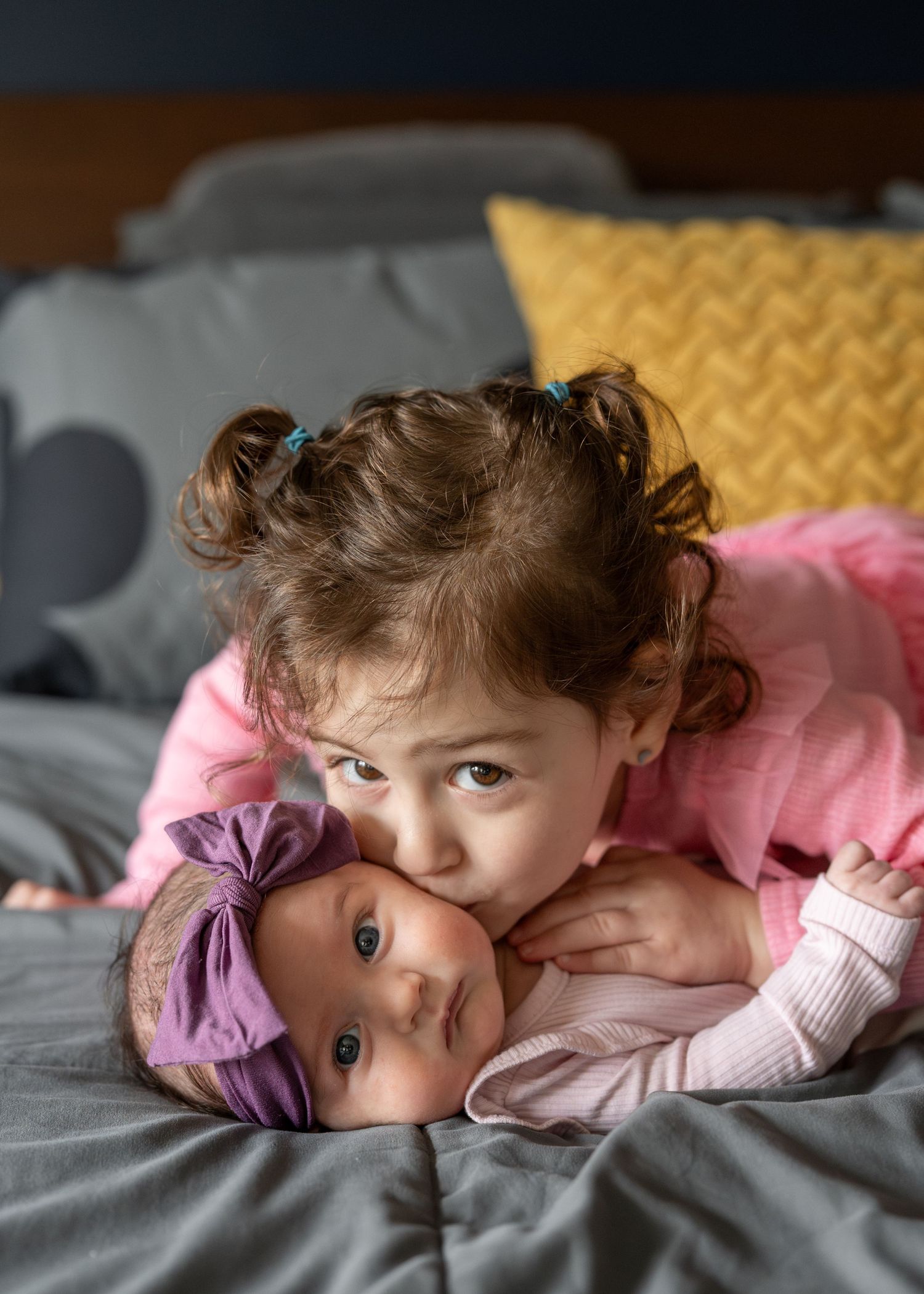 Baby in pink clothing lies on gray bedding while looking at camera with curious expression.