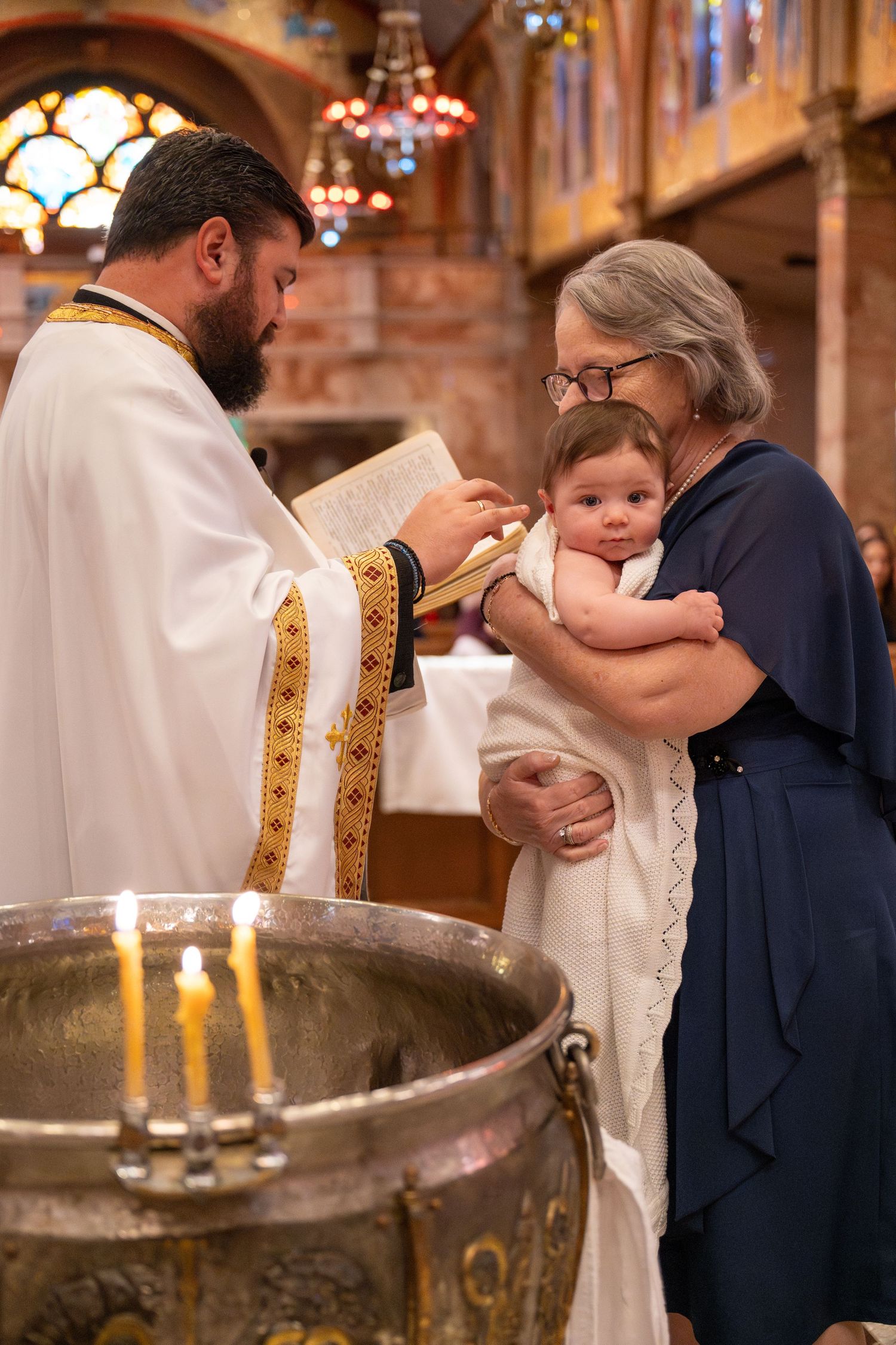 A religious baptism ceremony takes place in an ornate church with stained glass windows and candles.