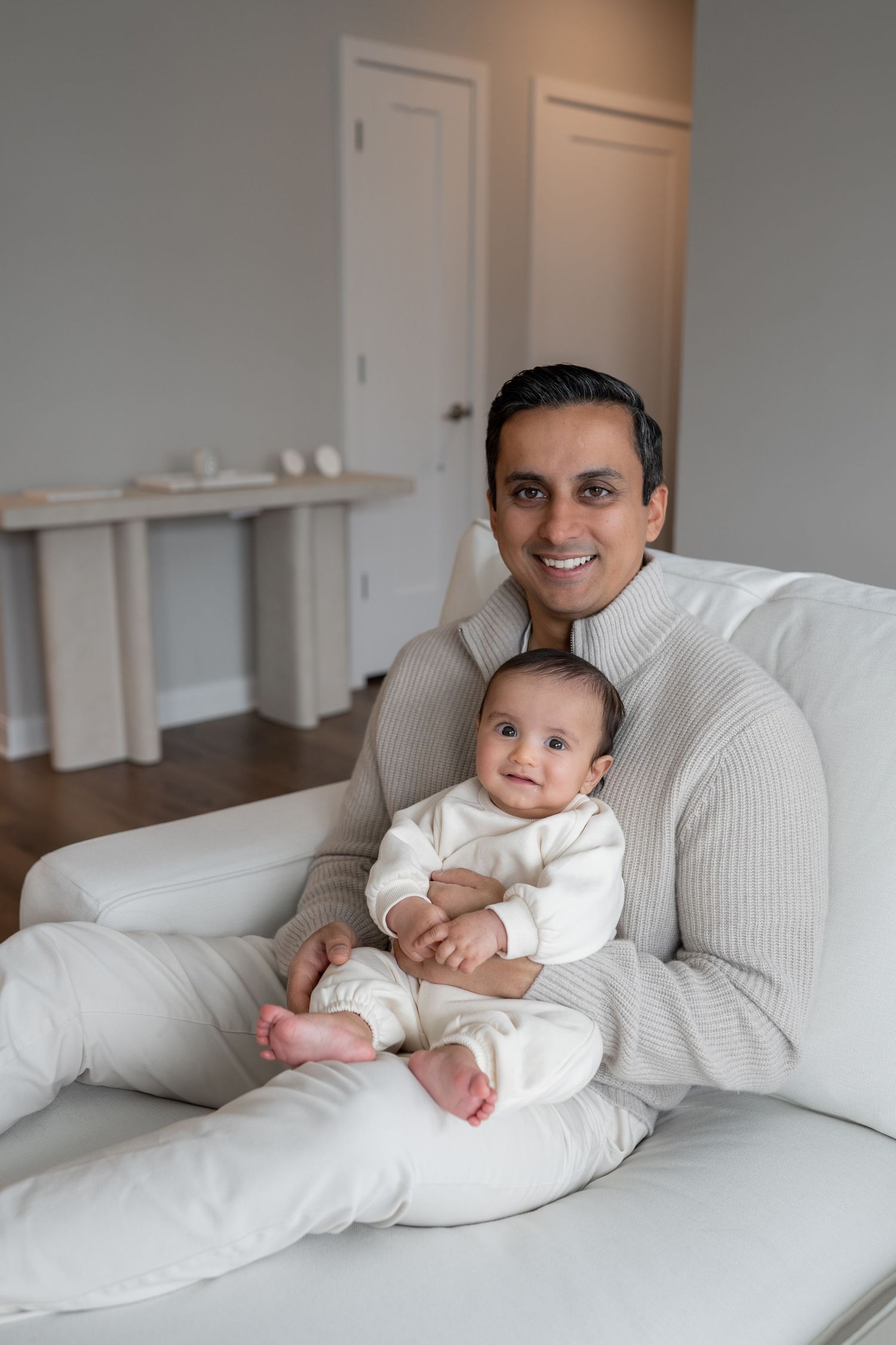 Smiling parent holding baby on white sofa in modern living room.
