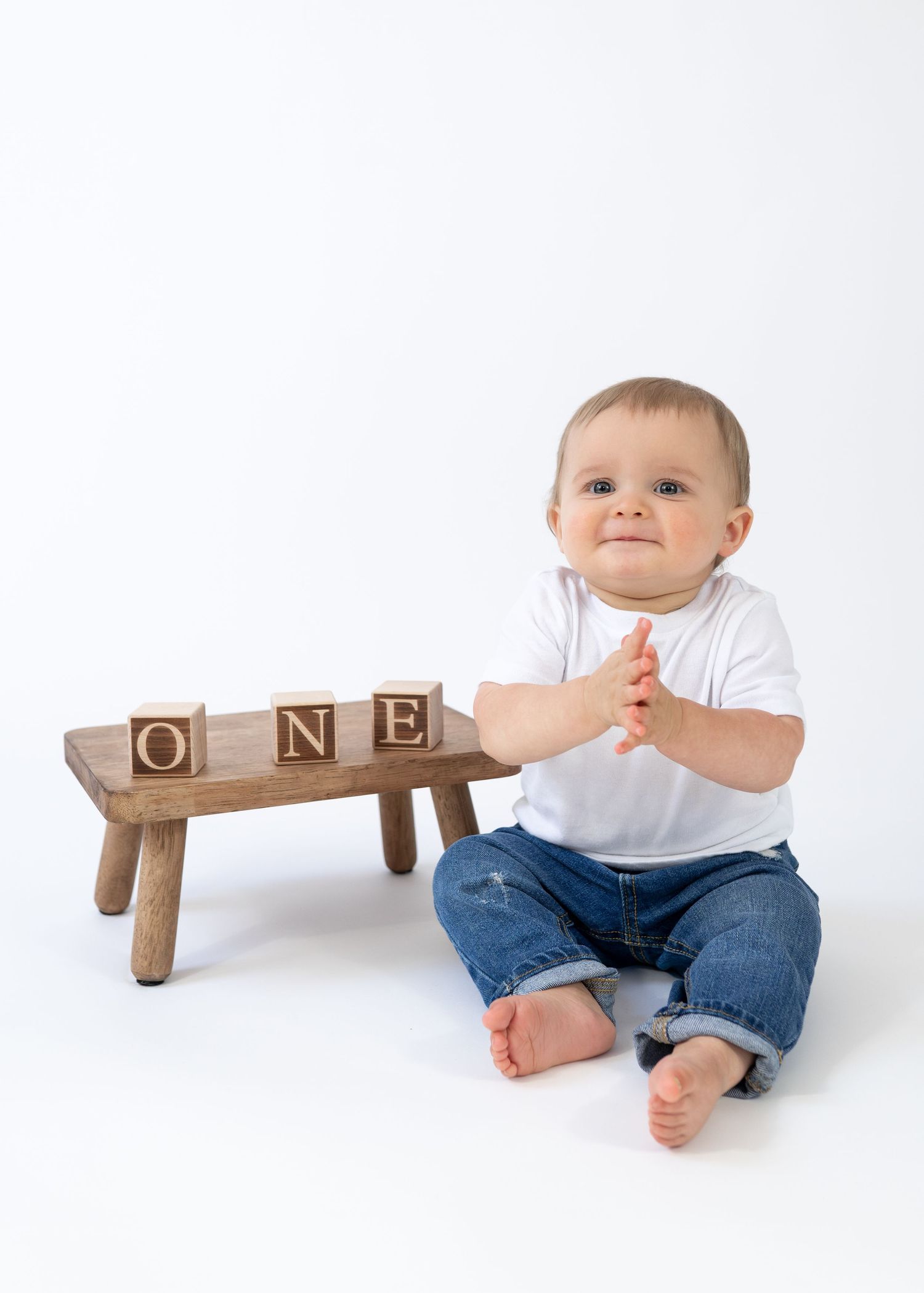 Adorable baby in white shirt and jeans sitting beside wooden blocks spelling ONE for first birthday photo.
