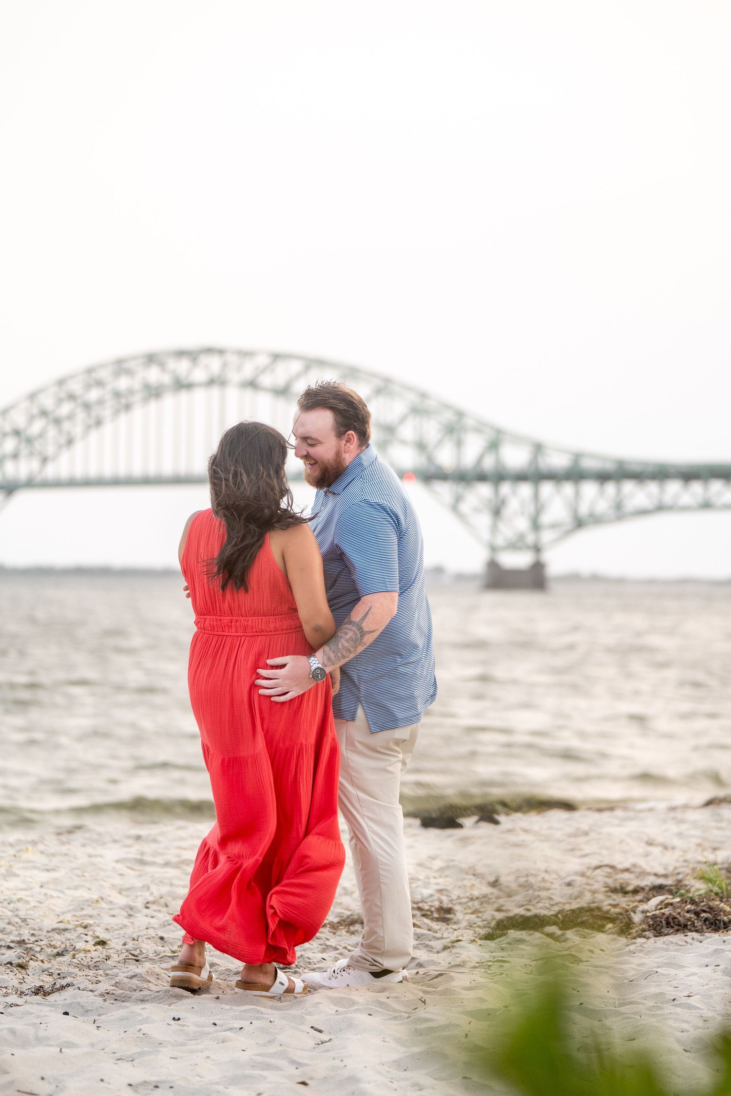 A couple embraces on a sandy beach with a large bridge structure visible across the water in the background.