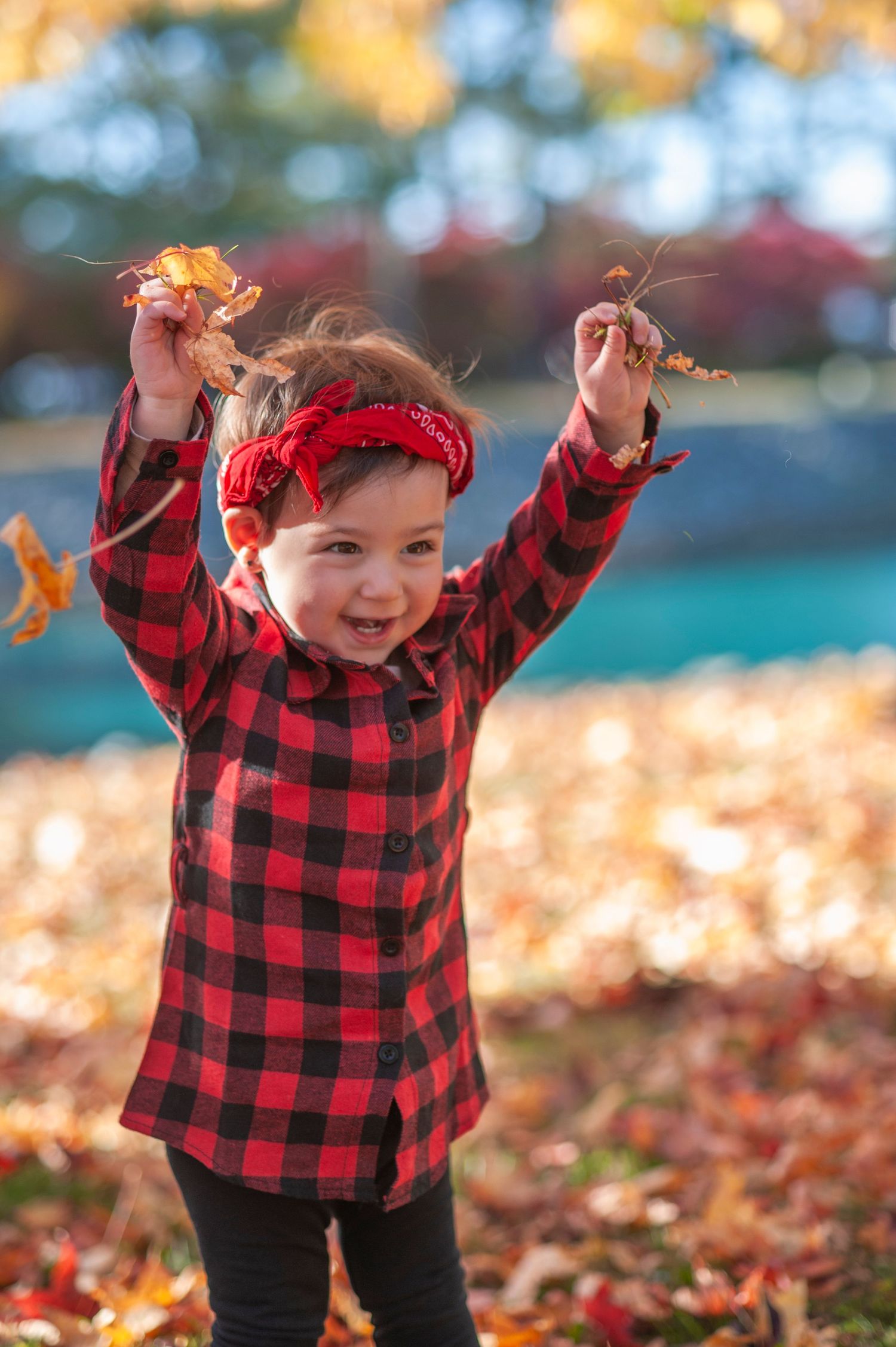 A child in a red buffalo plaid shirt plays joyfully with autumn leaves on a sunny fall day.