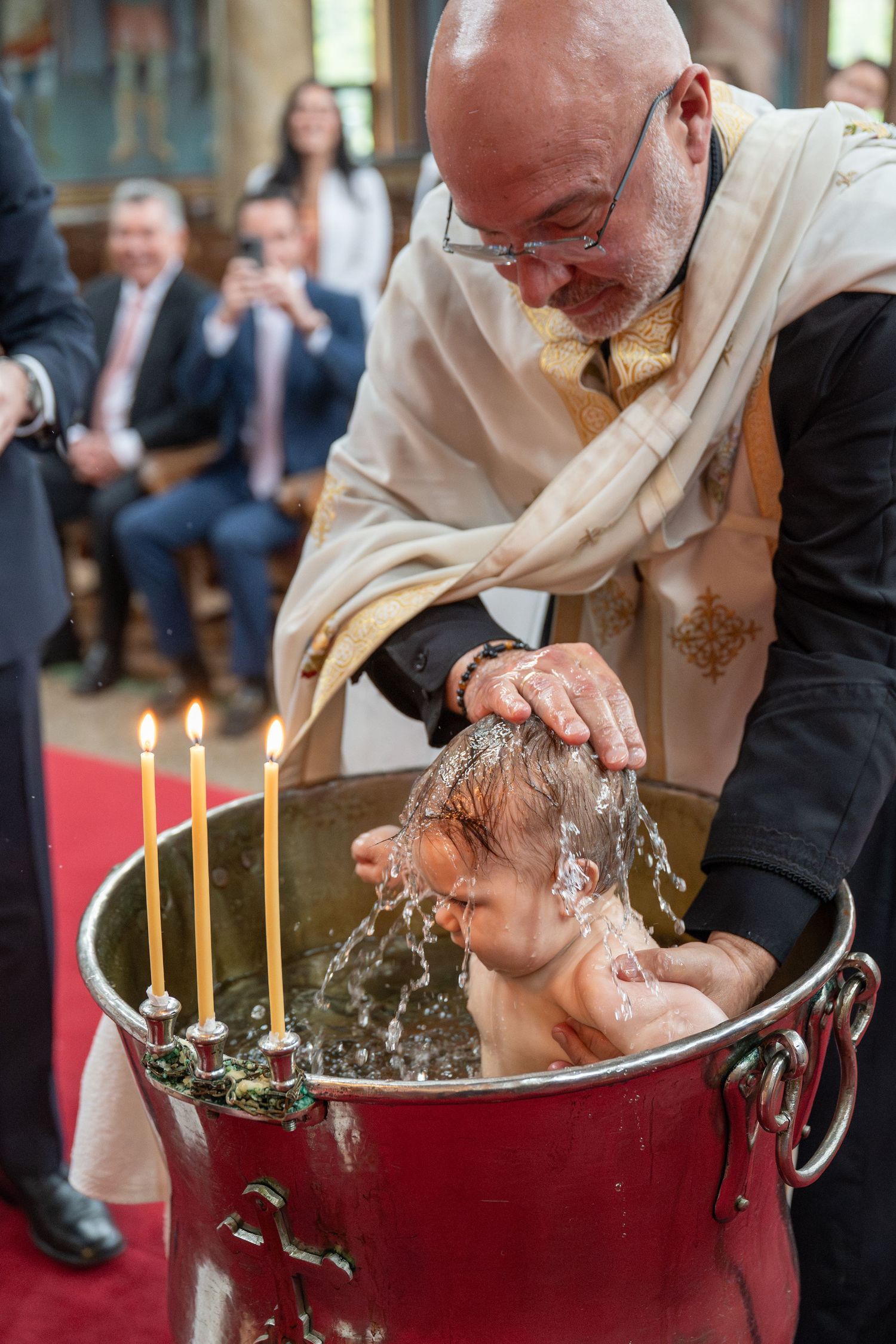 Priest performing a traditional Christian baptism, pouring water over an infant in a red baptismal font with candles.