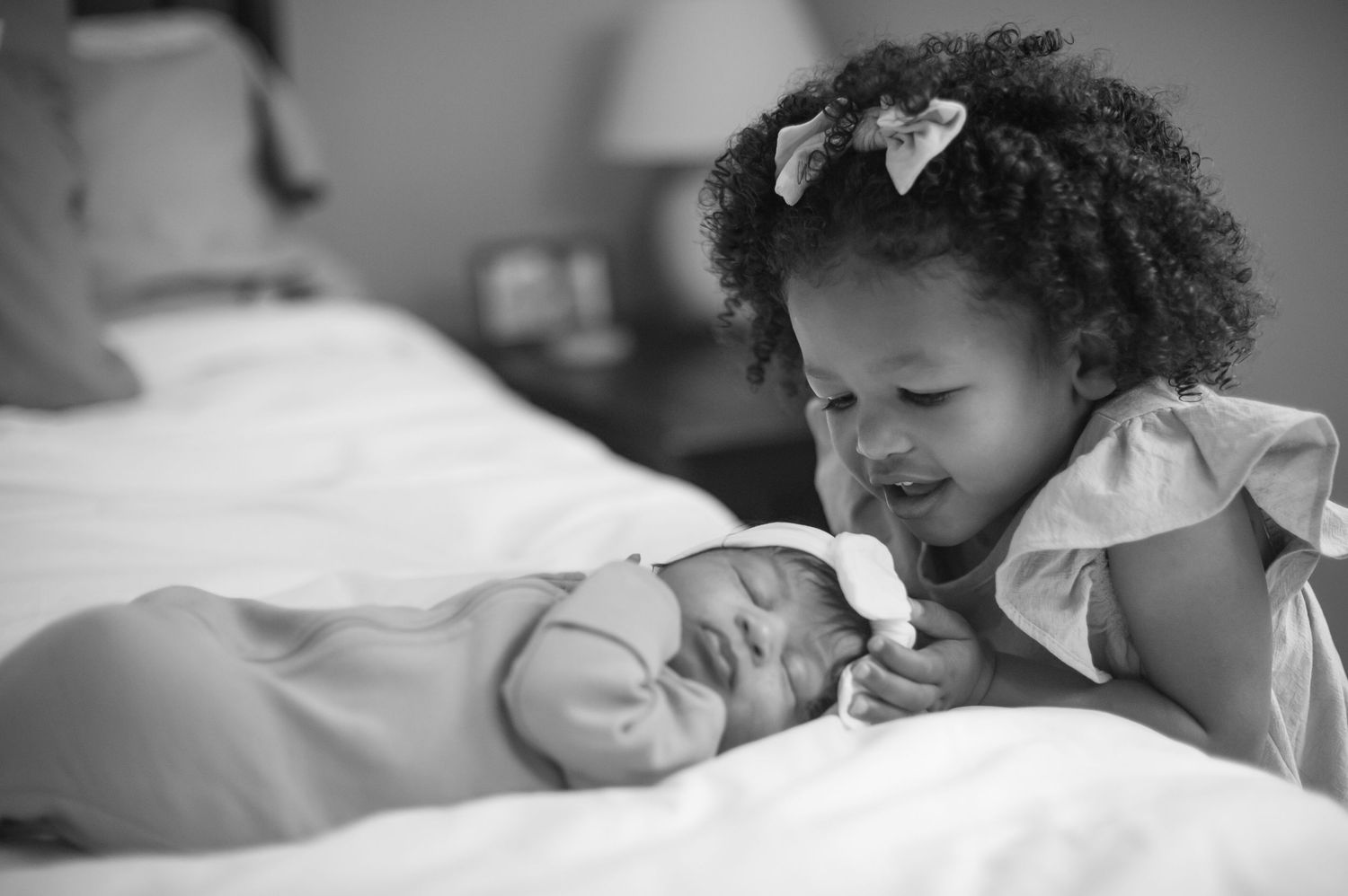 Black and white photo of a child leaning over to look at sleeping baby on bed.