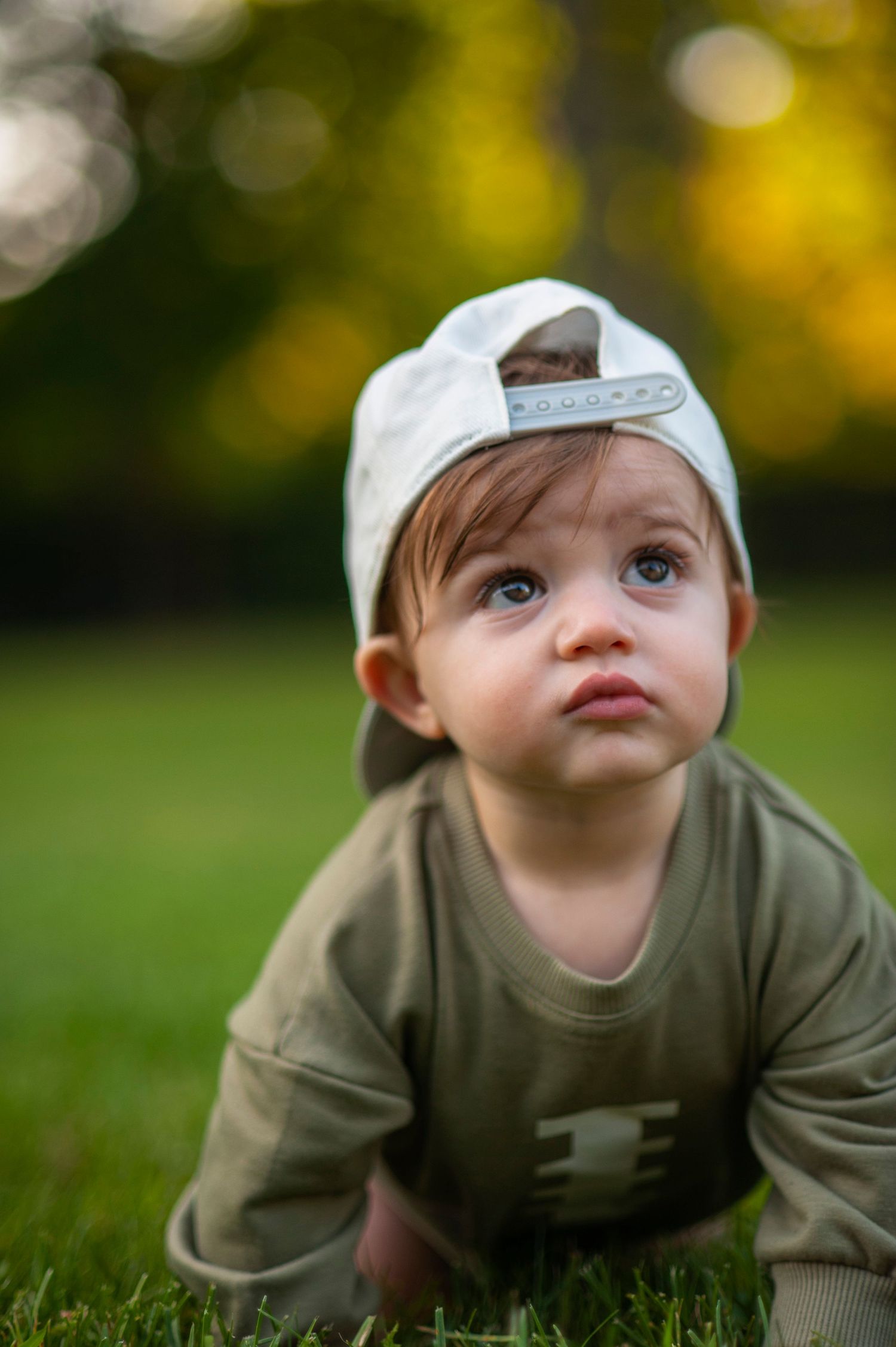 Baby in olive green shirt and white cap crawling on grass with bokeh background.