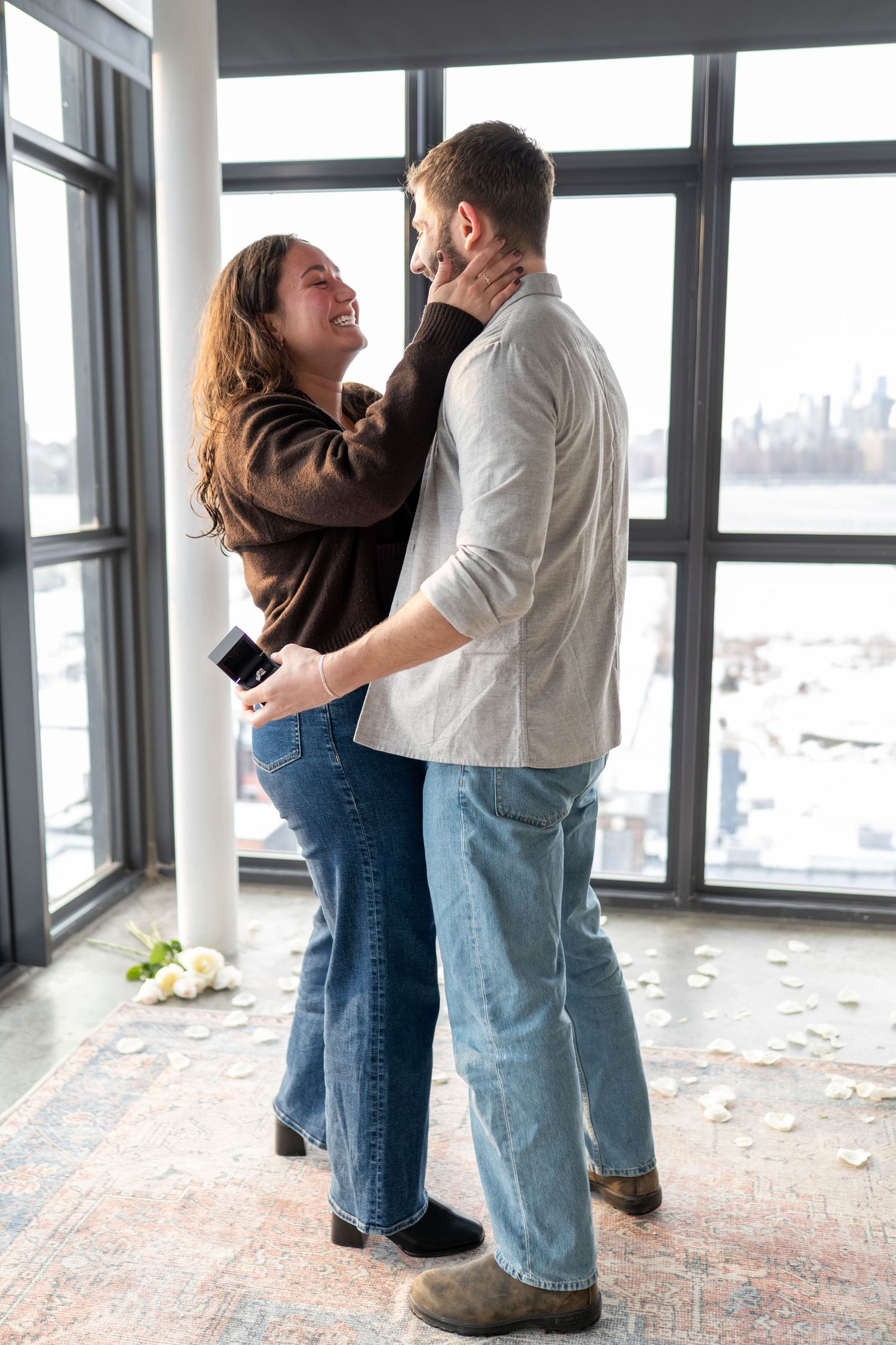 A couple embraces near large windows in an indoor space with rose petals scattered on the floor.