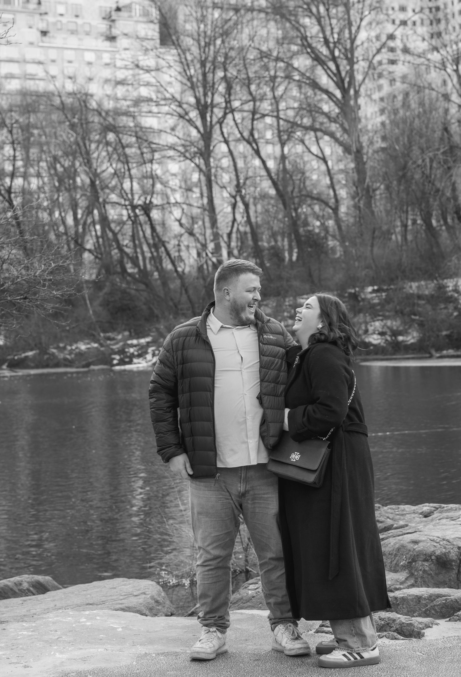 A couple shares an intimate moment on snowy rocks by a lake with city buildings in the background.