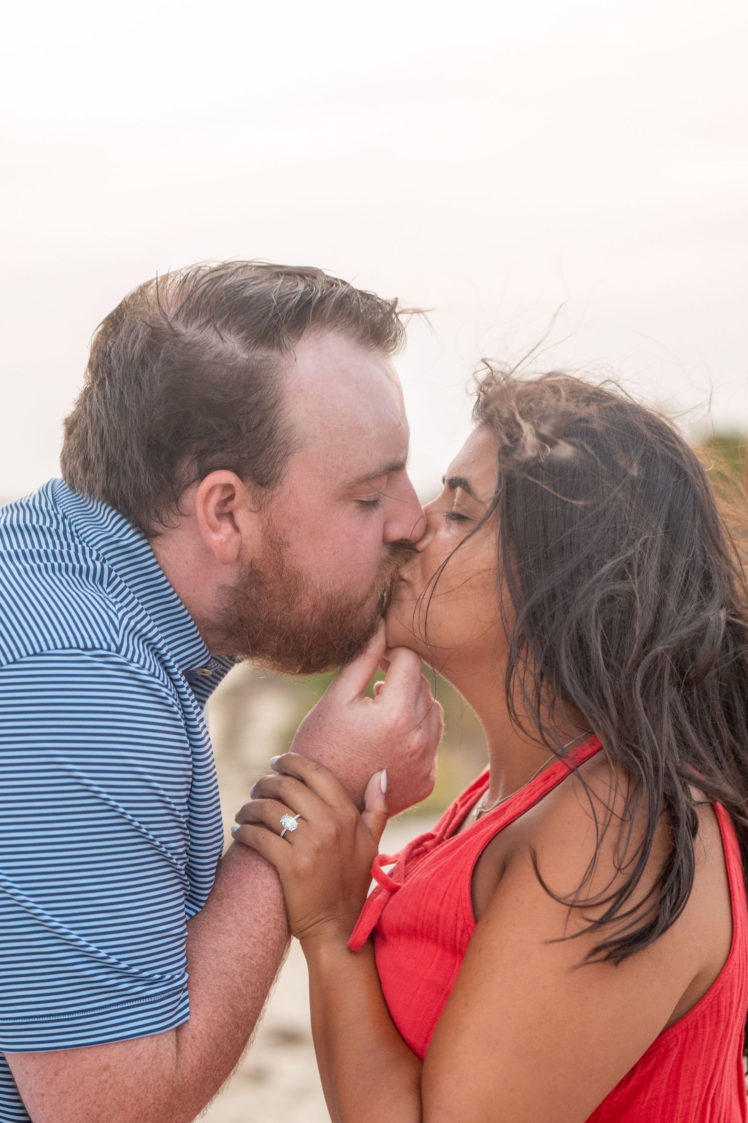 A couple in a blue striped shirt and red top share a romantic kiss on a sunny beach.