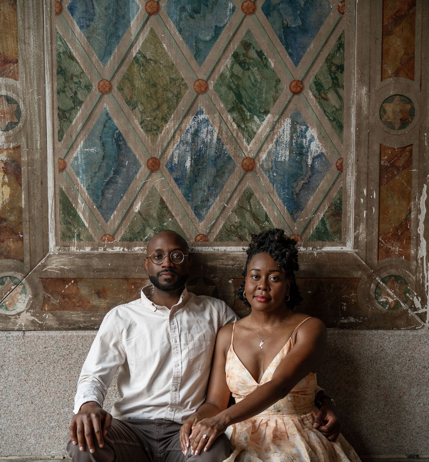 A couple sits together against a decorative wall with diamond patterns and frescoes in muted tones.