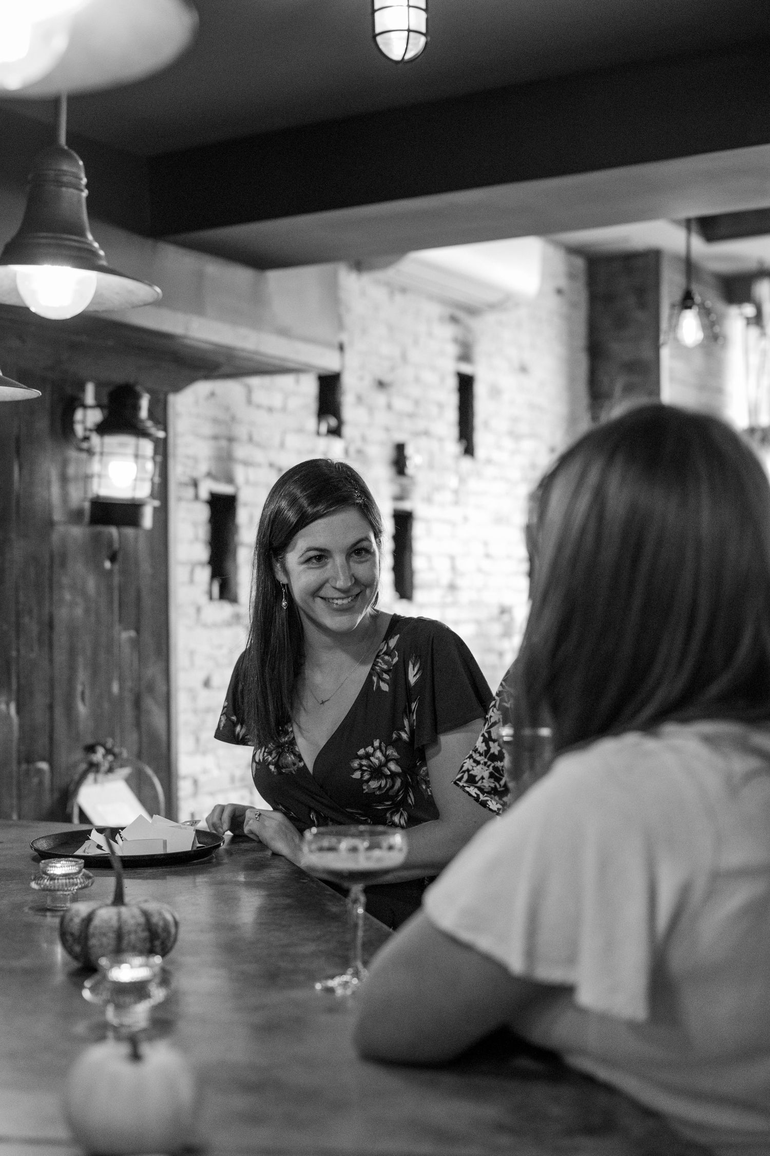 Black and white photograph of people conversing at a bar with industrial lighting and brick walls.