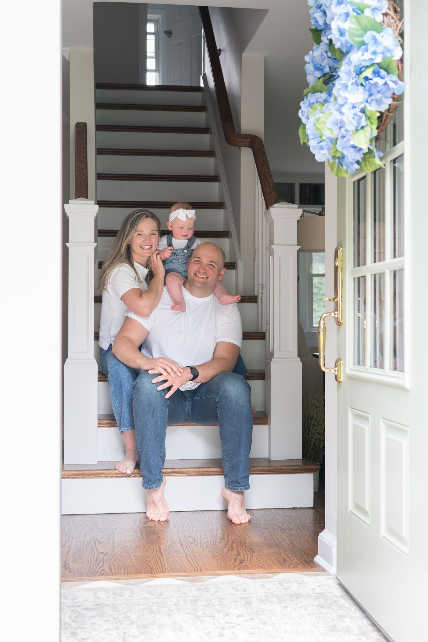 A family sits together on white wooden stairs in an entryway with hardwood floors and blue hydrangeas by the door.