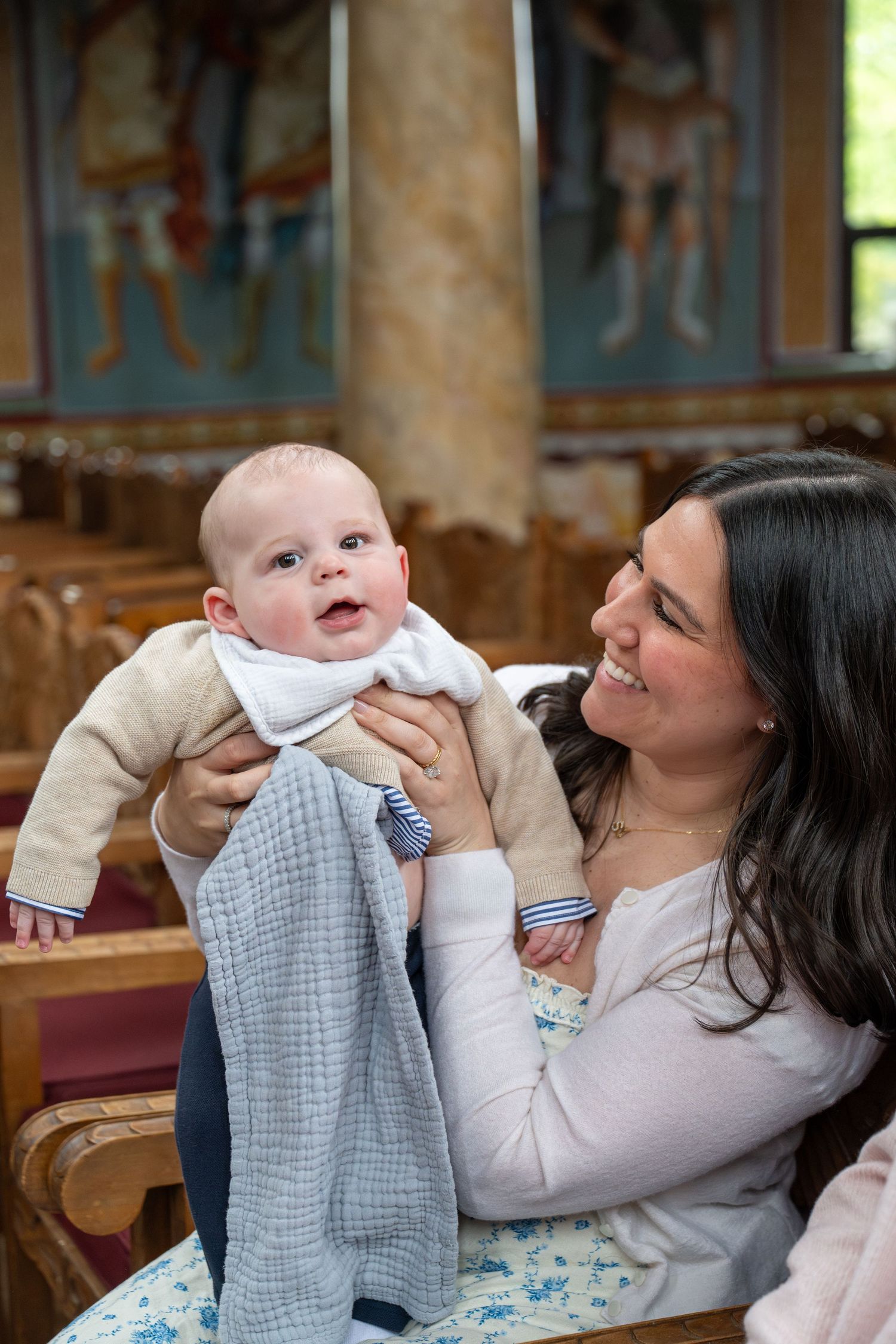 A mother holds her smiling baby during a baptism ceremony in a church with ornate decorations visible in the background.