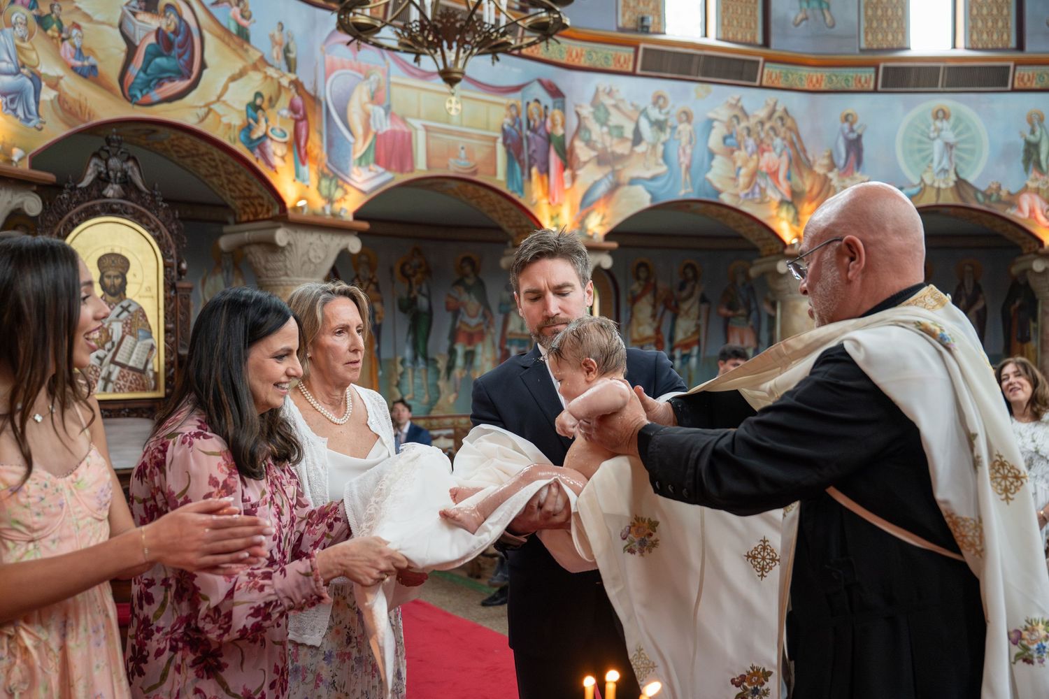 Orthodox baptism ceremony with priest, parents, and godparents holding infant in ornate church with colorful frescoes.