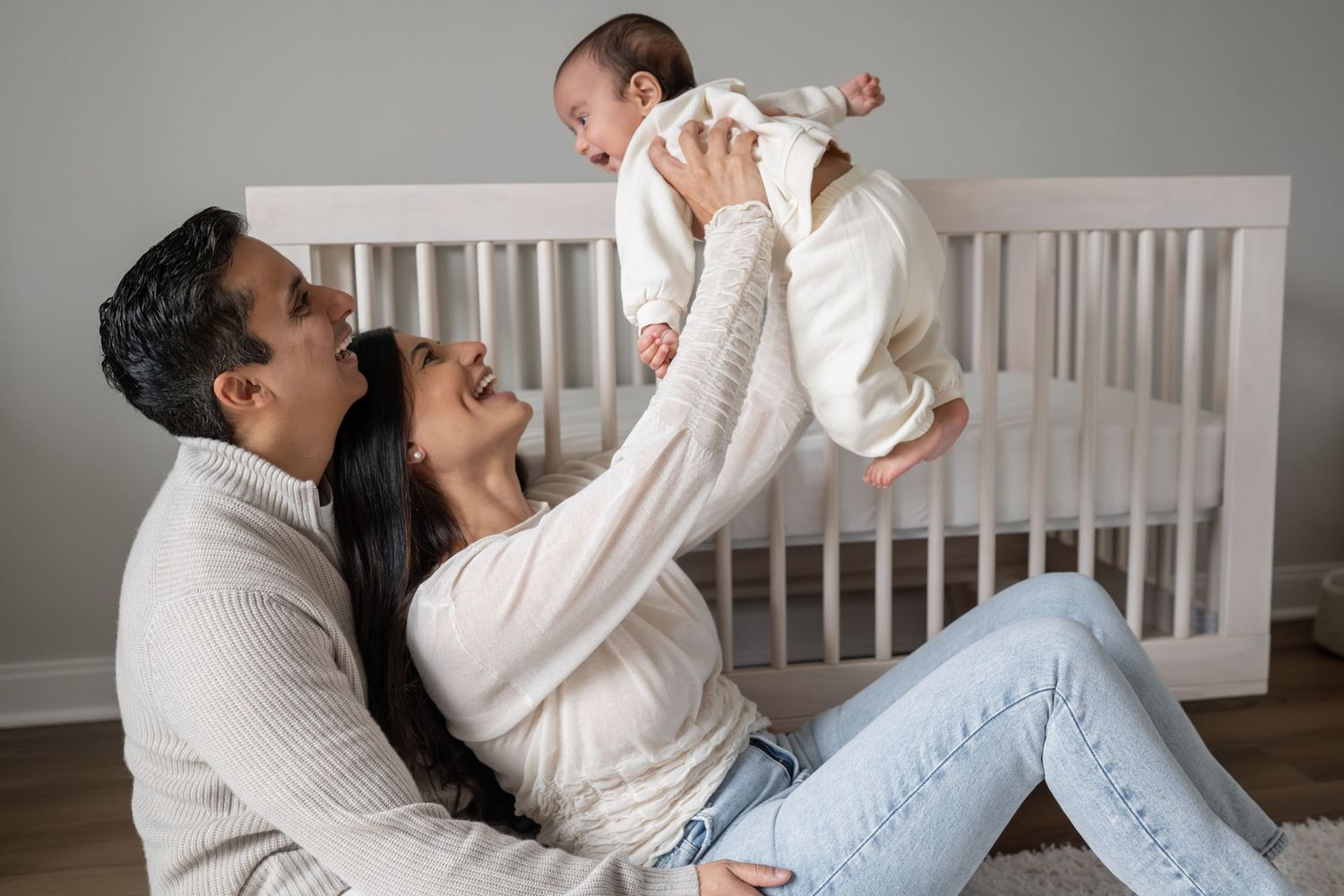 Joyful parents sitting on floor lifting laughing baby in white onesie in front of white nursery crib.