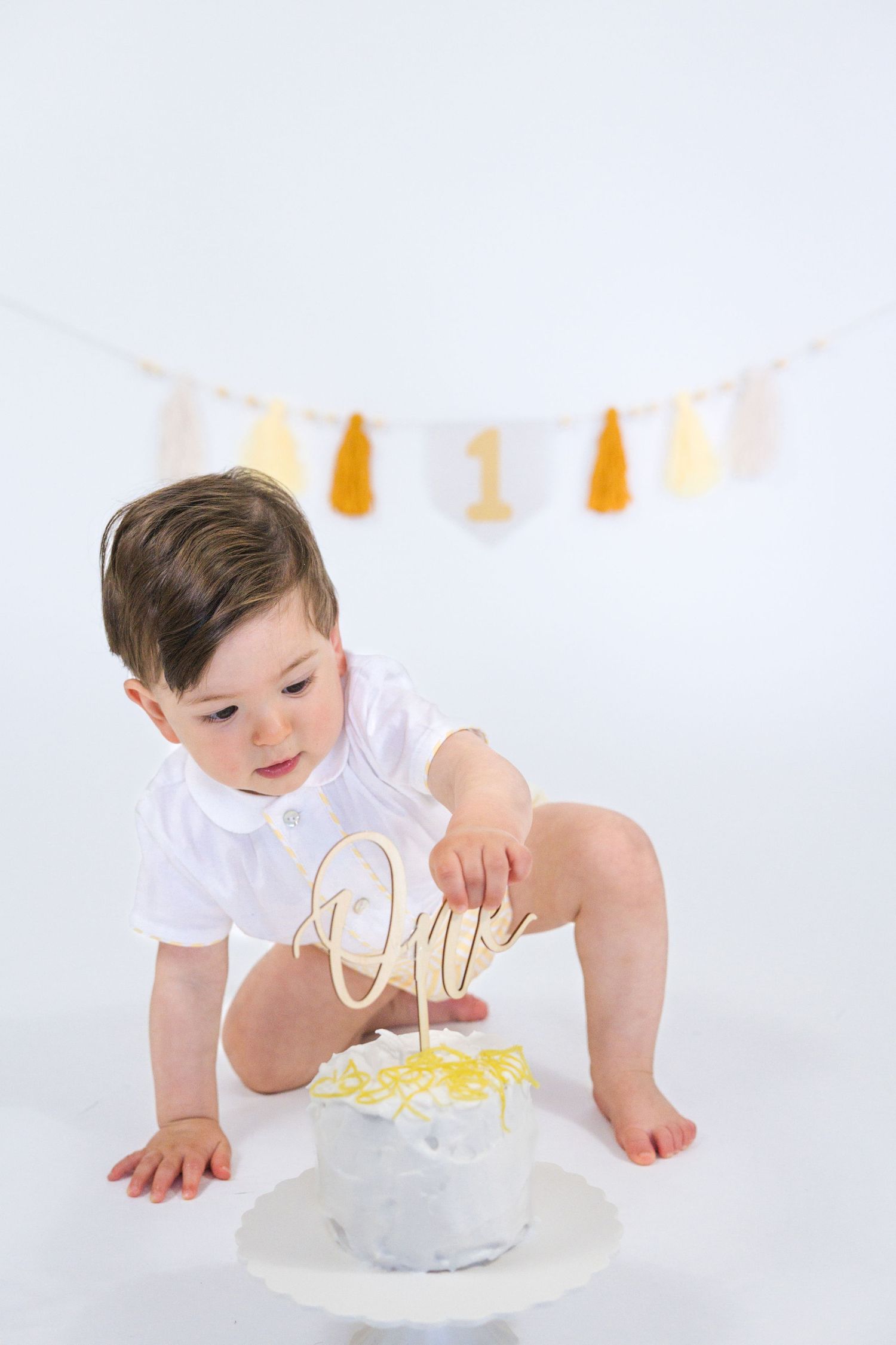 Baby in white outfit explores birthday cake on white background with yellow banner.