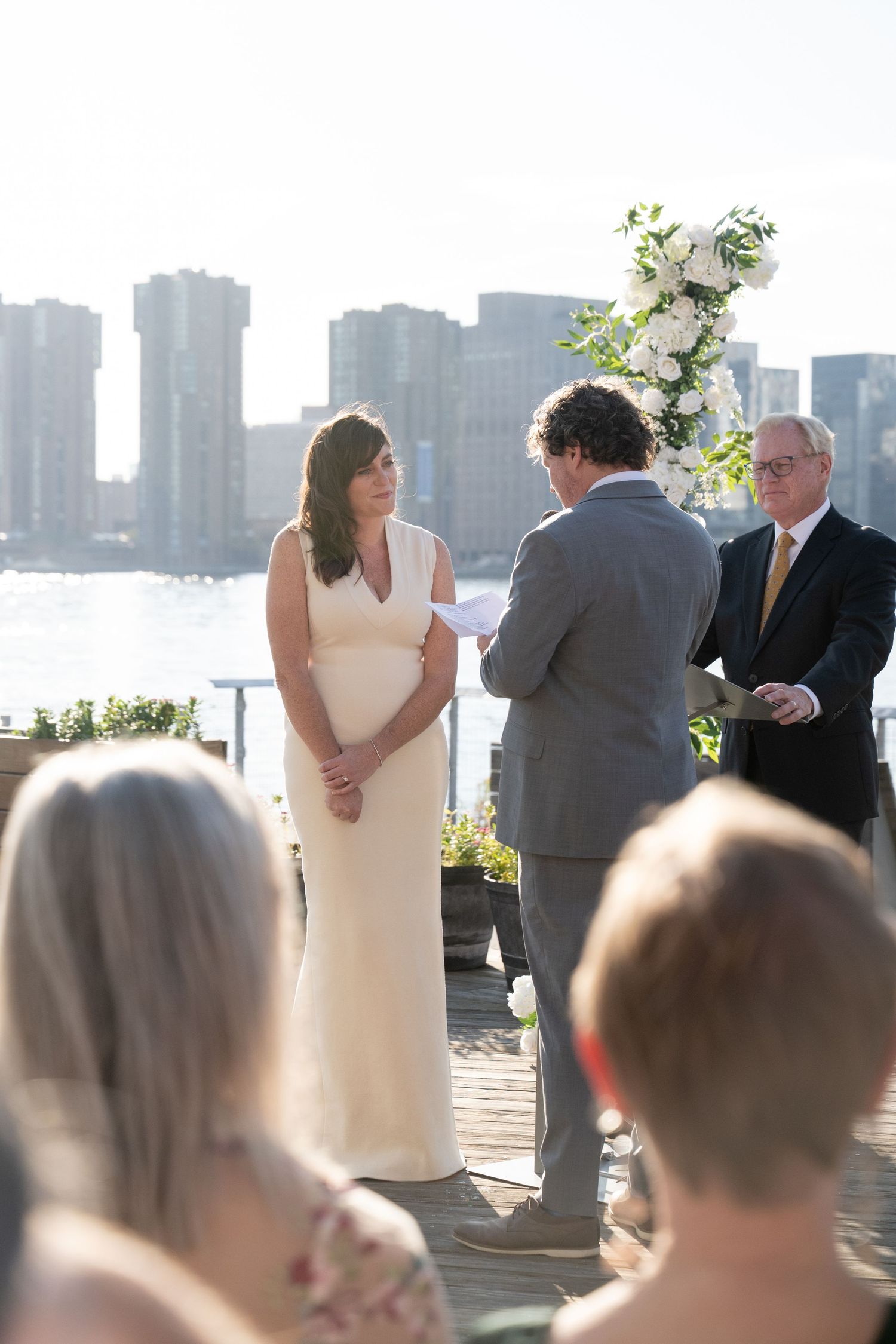 An outdoor wedding ceremony takes place on a waterfront deck with tall city buildings visible across the water.