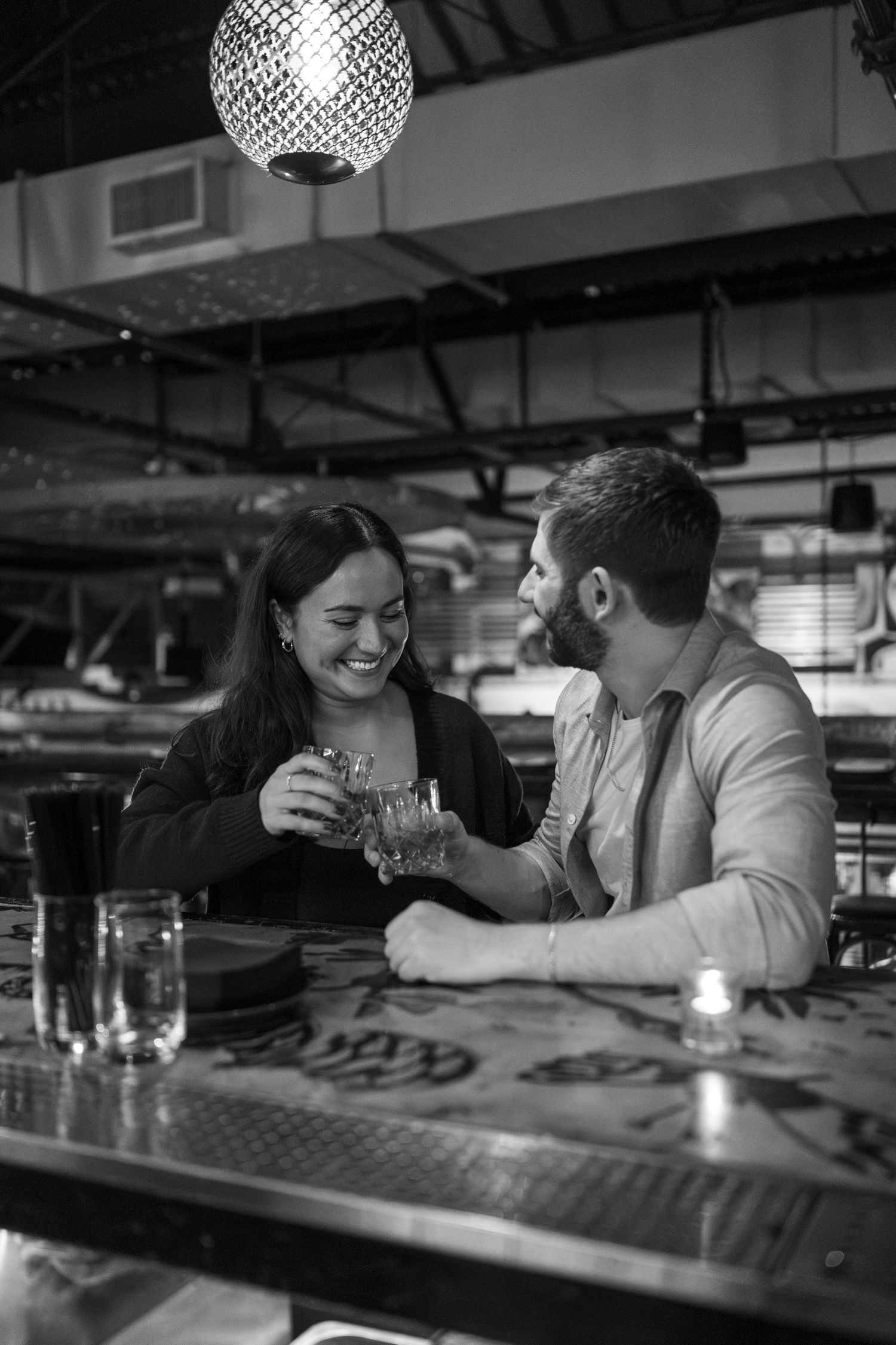 A couple sharing intimate moments and laughter at a bar counter under decorative lighting.