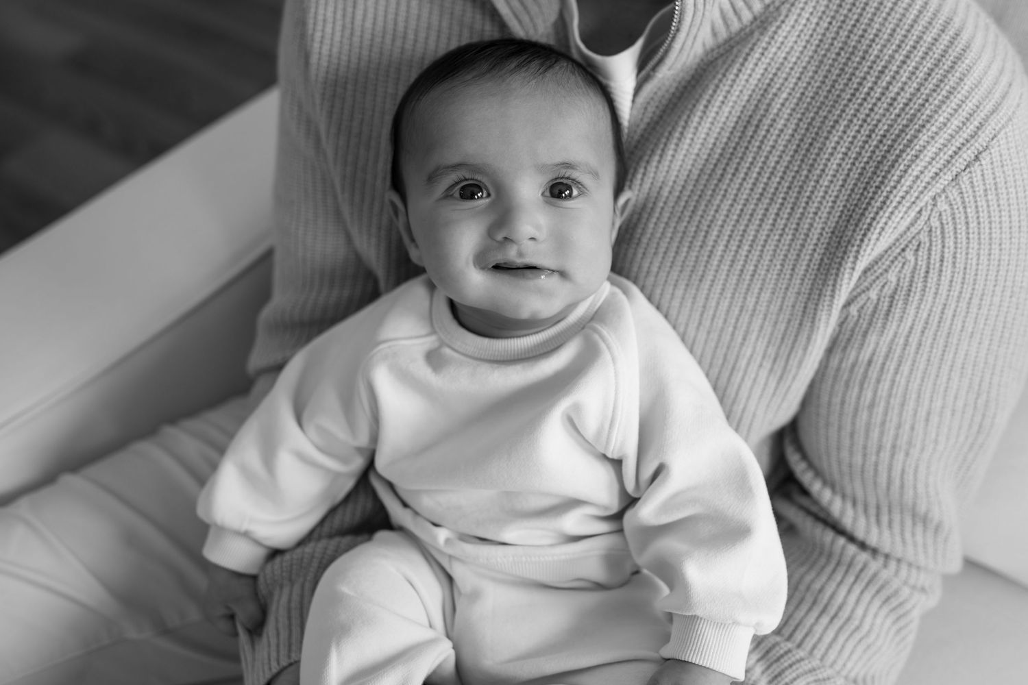 Adorable baby in white outfit sitting on parent's lap, black and white photo.