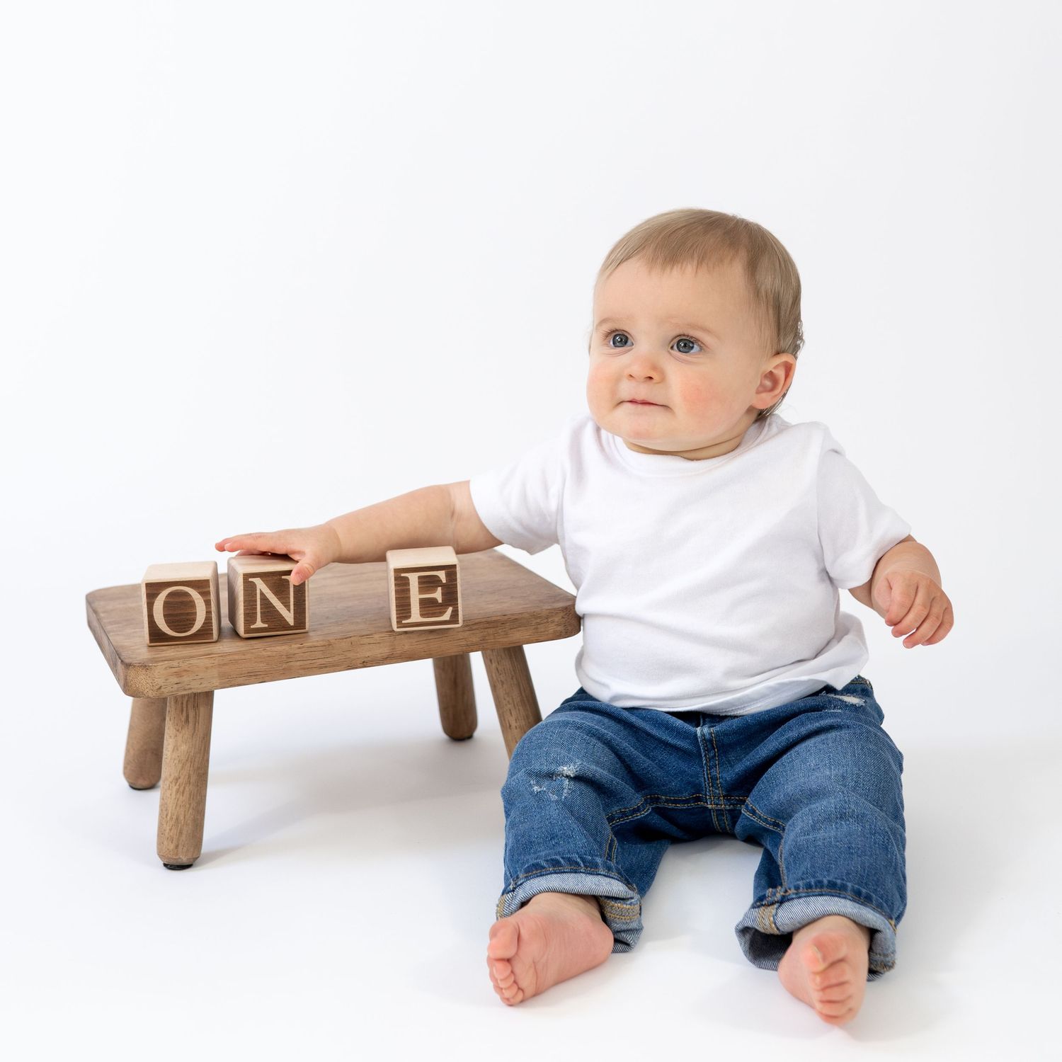 Baby in white onesie and jeans sitting beside wooden stool with ONE alphabet blocks, first birthday photo.