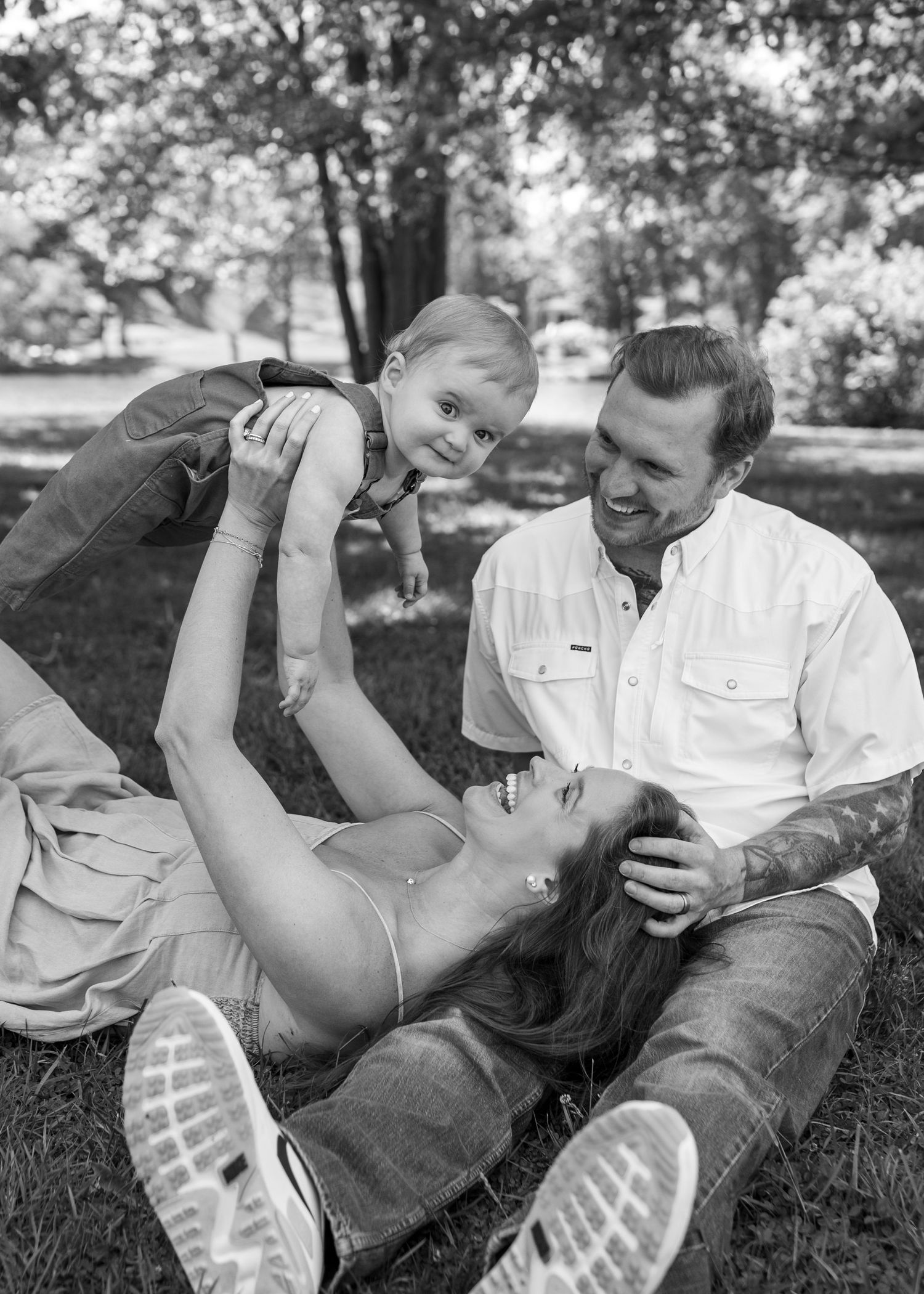 Black and white photo of a happy family playing in the park, mother lifting baby while father smiles nearby.