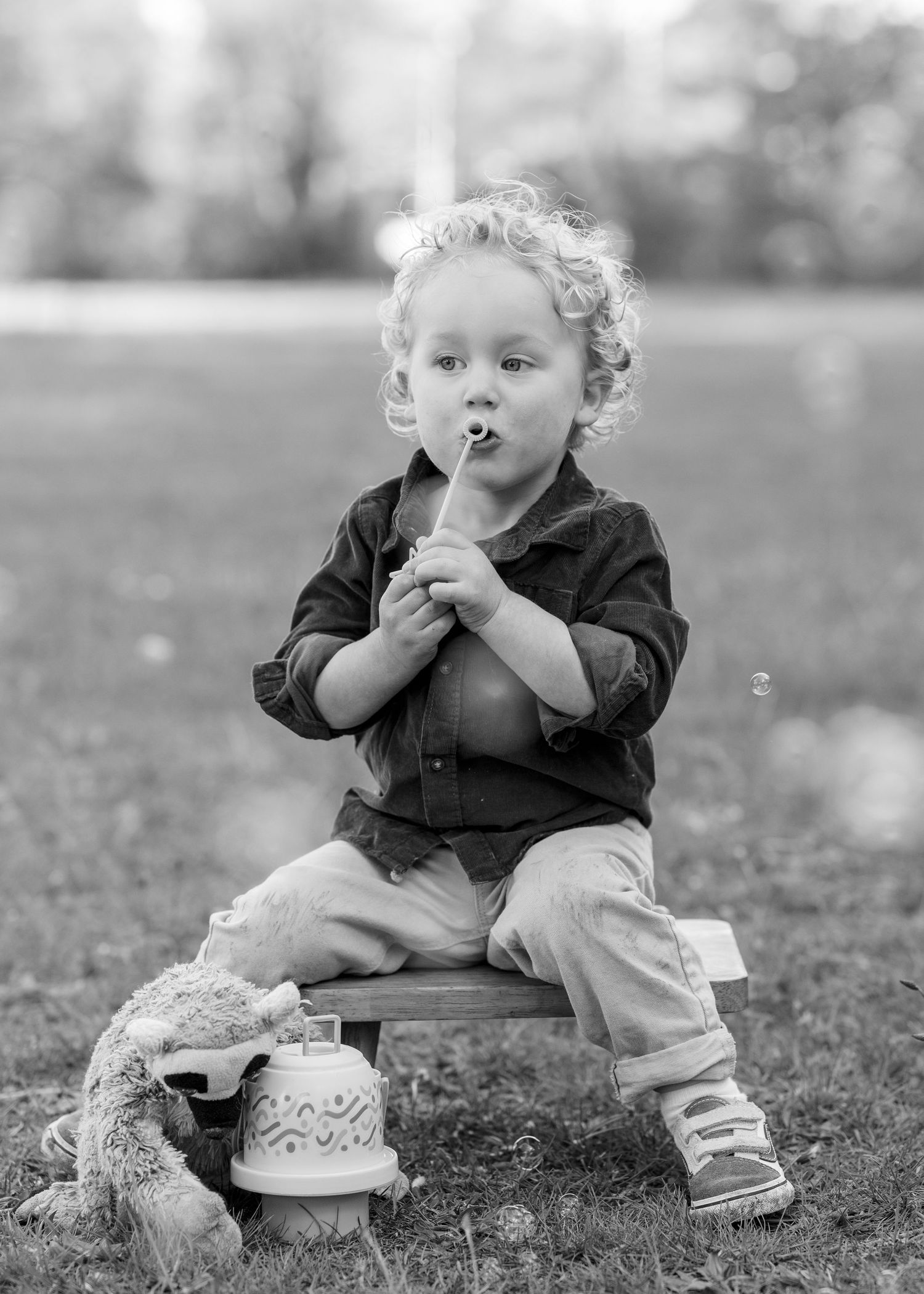 Cute curly-haired toddler blowing bubbles while sitting outdoors in black and white photo.