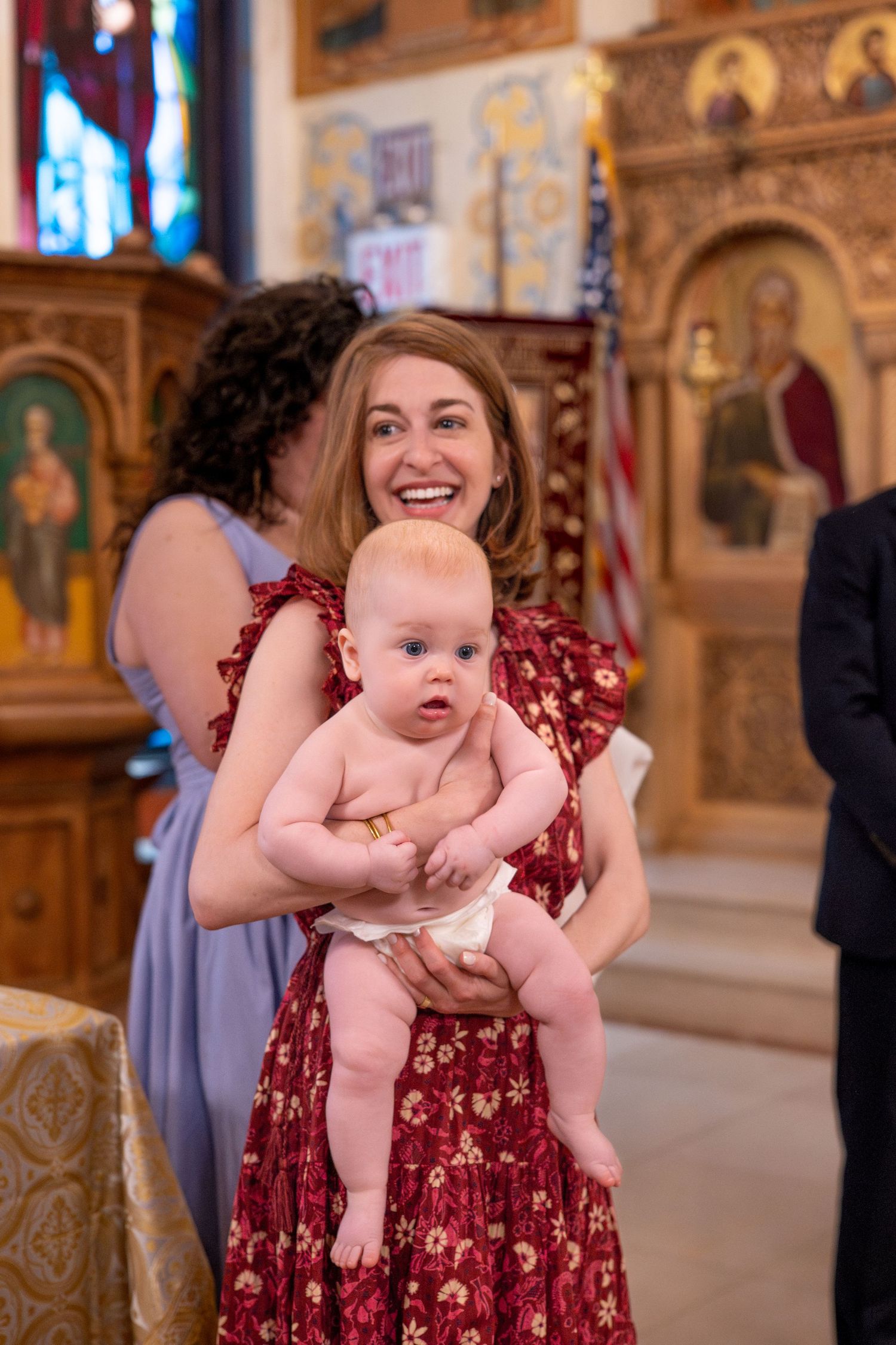 A baby in pink clothing is held up during a church ceremony with ornate decorations visible in the background.