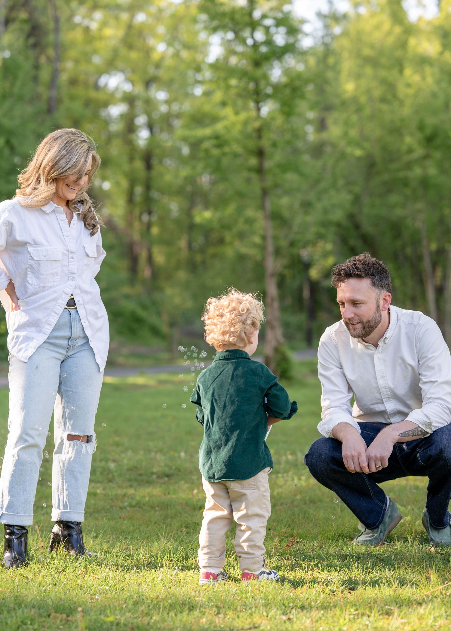Young family in green park, mother and father in white shirts watching their curly-haired toddler in green shirt.