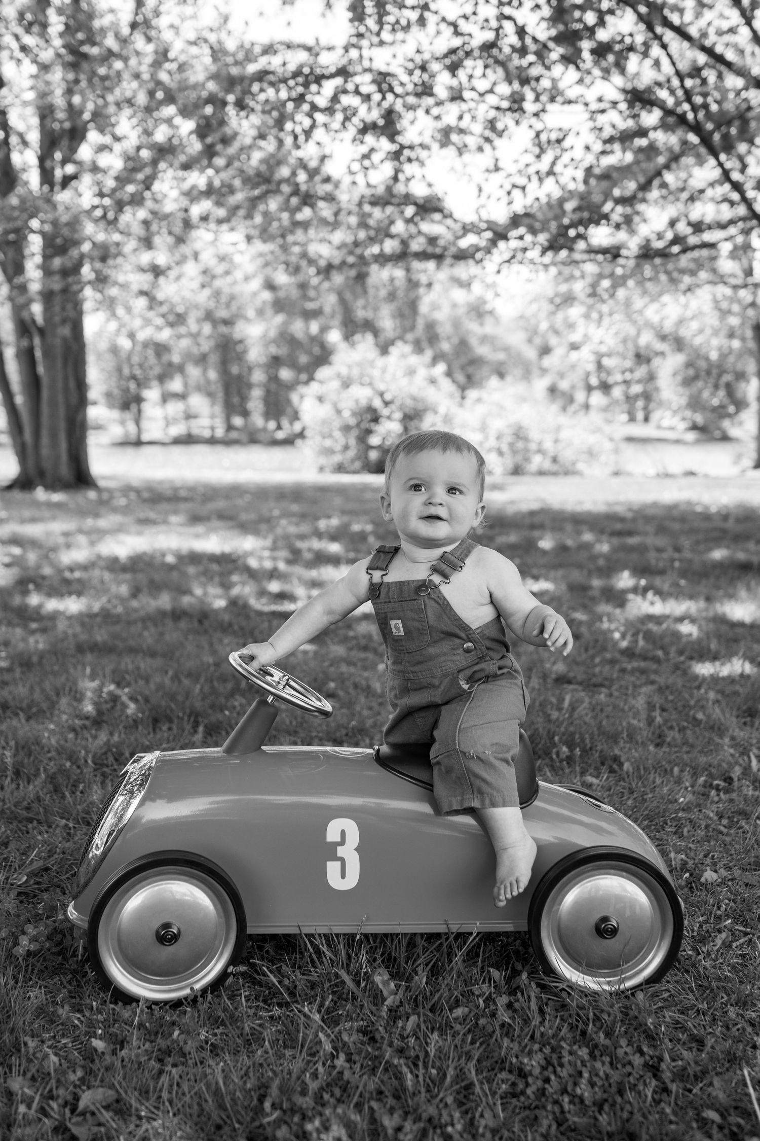 Black and white photo of a toddler boy sitting in a vintage pedal car numbered 3 on green grass in a park.