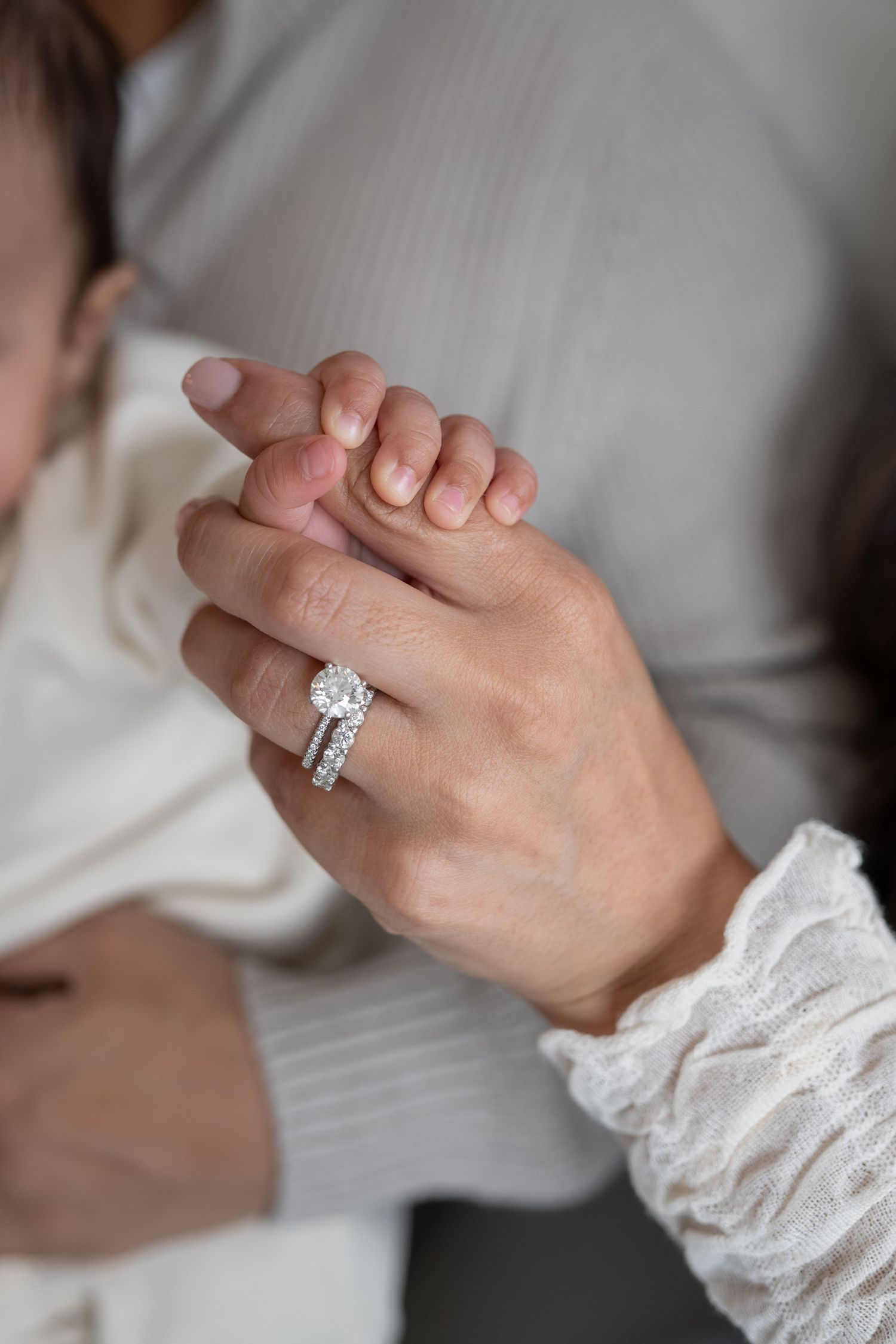 Close-up of a woman's hand wearing a diamond halo engagement ring and wedding band, gently holding a newborn baby's tiny fingers.