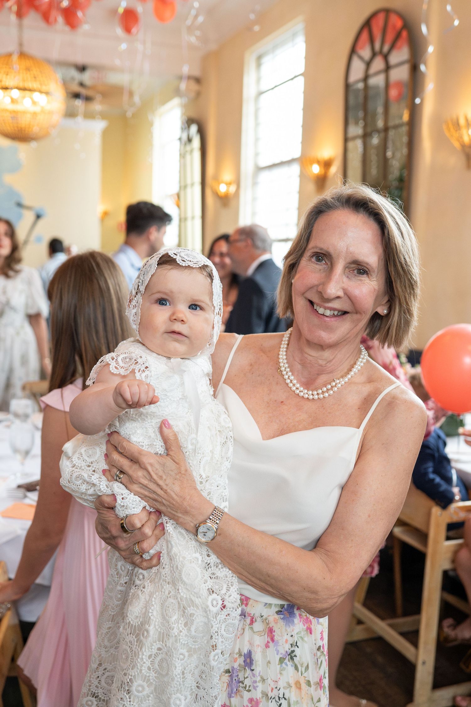 Smiling grandmother holding baby in white lace christening gown at baptism party.