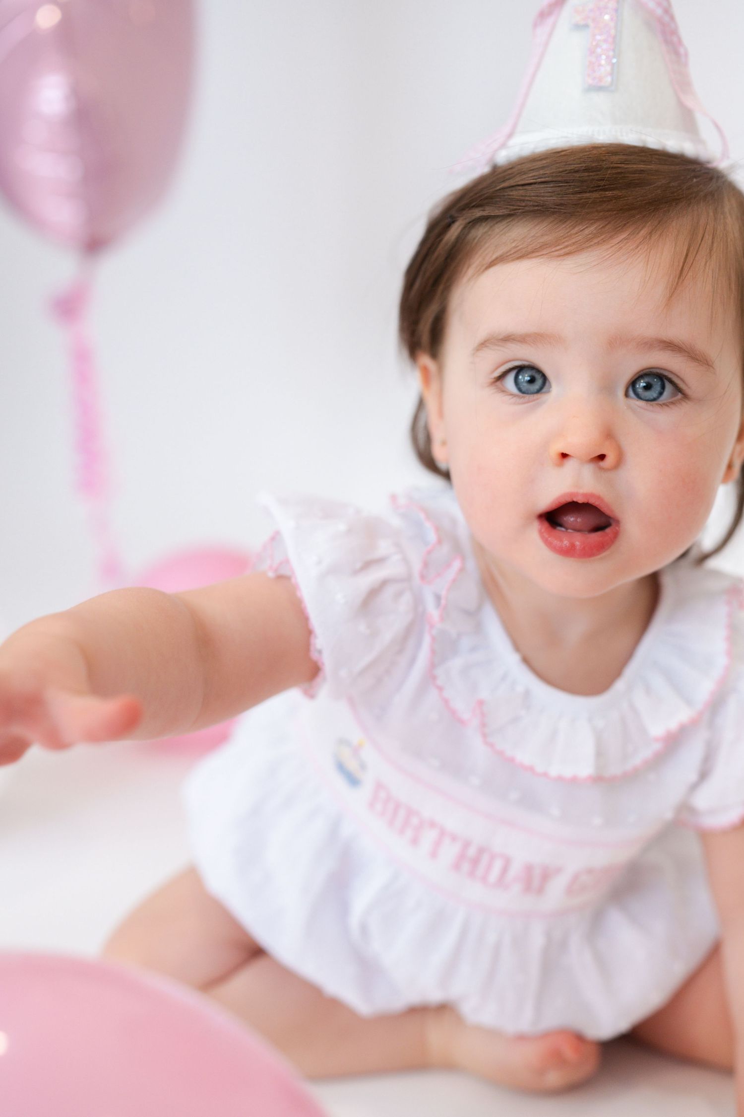 Baby in white dress with pink accents playing with balloons during photo shoot.