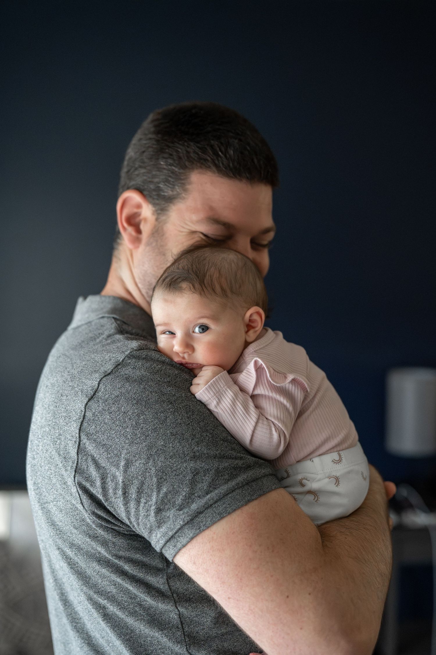 Baby in light pink outfit being held and comforted against gray shirt in dark room.