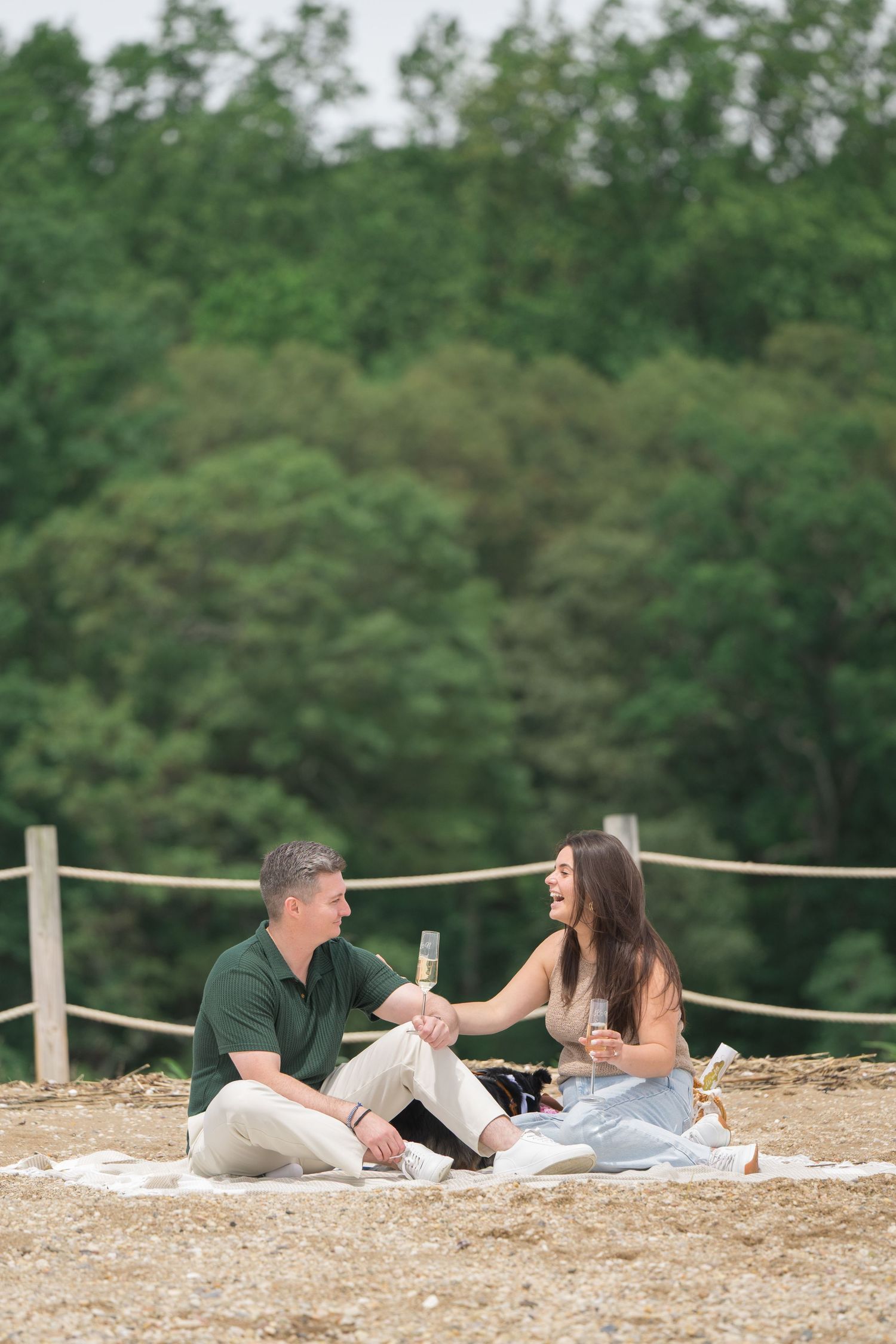A couple lying on a sandy beach sharing champagne and celebrating their engagement surrounded by trees.