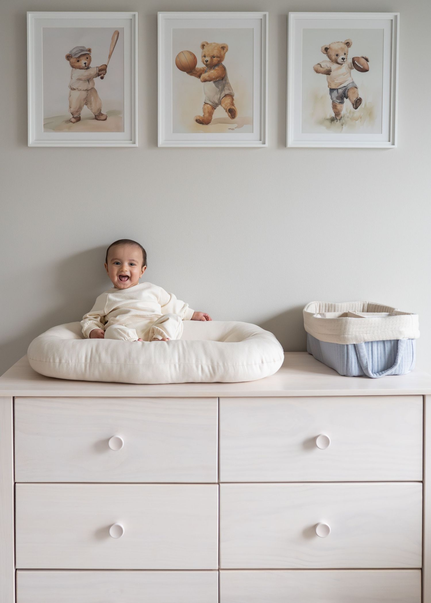 Laughing baby in white onesie on changing pad atop wooden dresser with teddy bear sports art prints on wall.