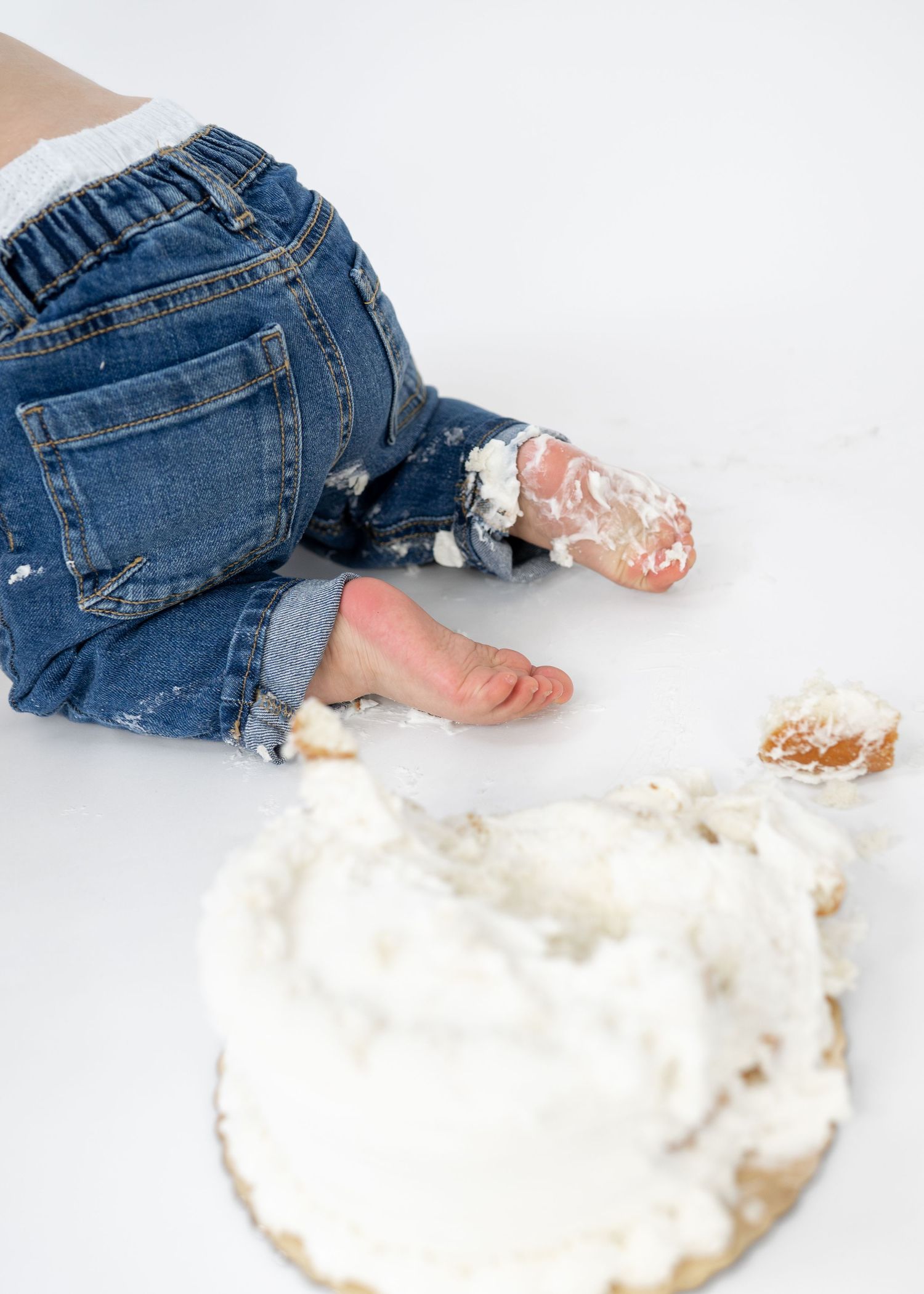 Baby in jeans smashing a white frosted cake with bare feet during a cake smash photo session.