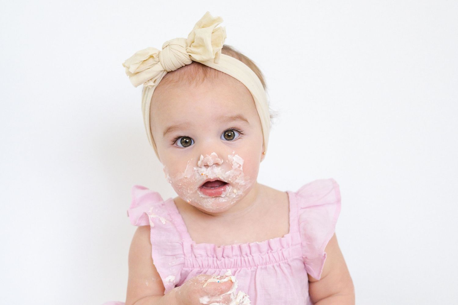 Baby in pink ruffled dress and cream bow headband smiling during cake smash photoshoot.