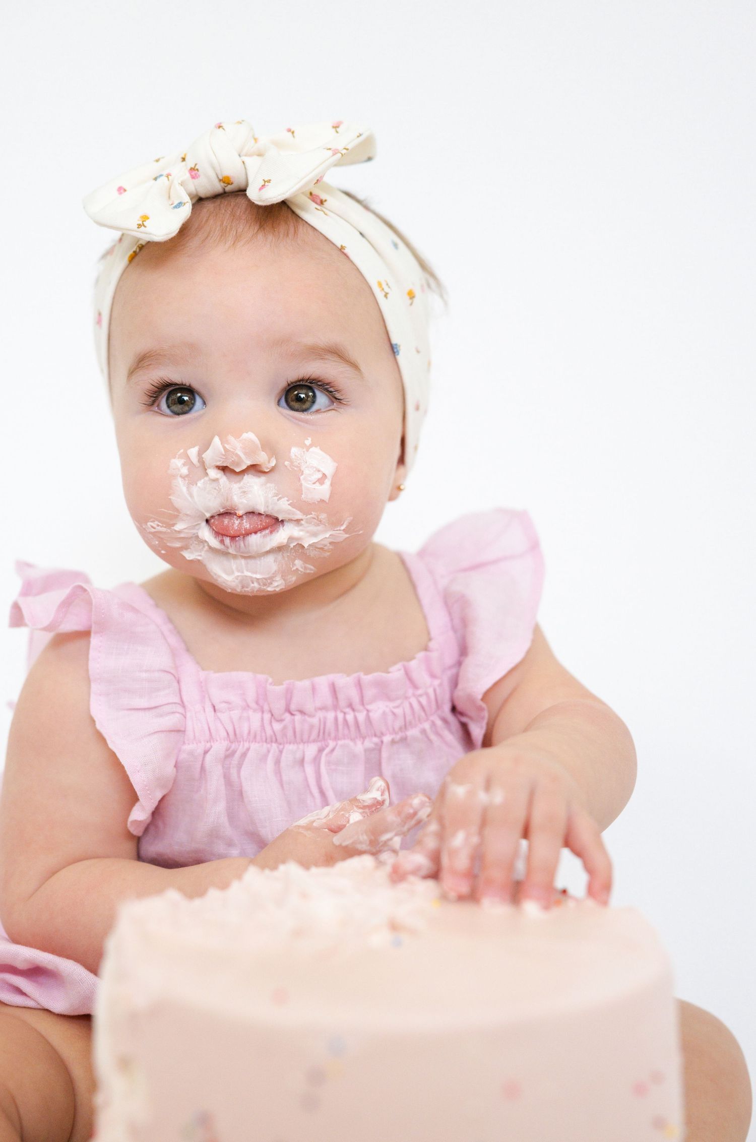 Baby in pink dress with messy face enjoying birthday cake smash photoshoot.