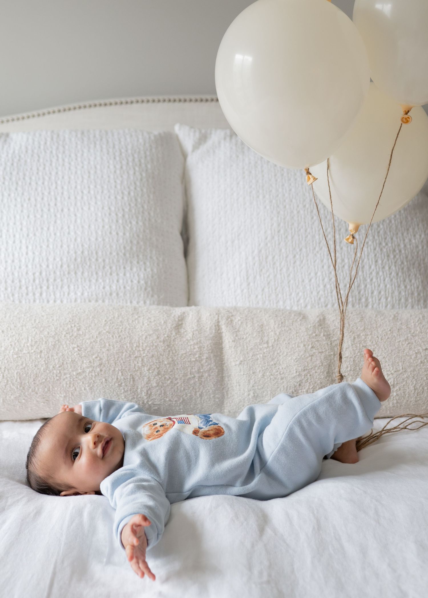 Adorable baby in blue onesie lies on white bed next to cream balloons on a stick.