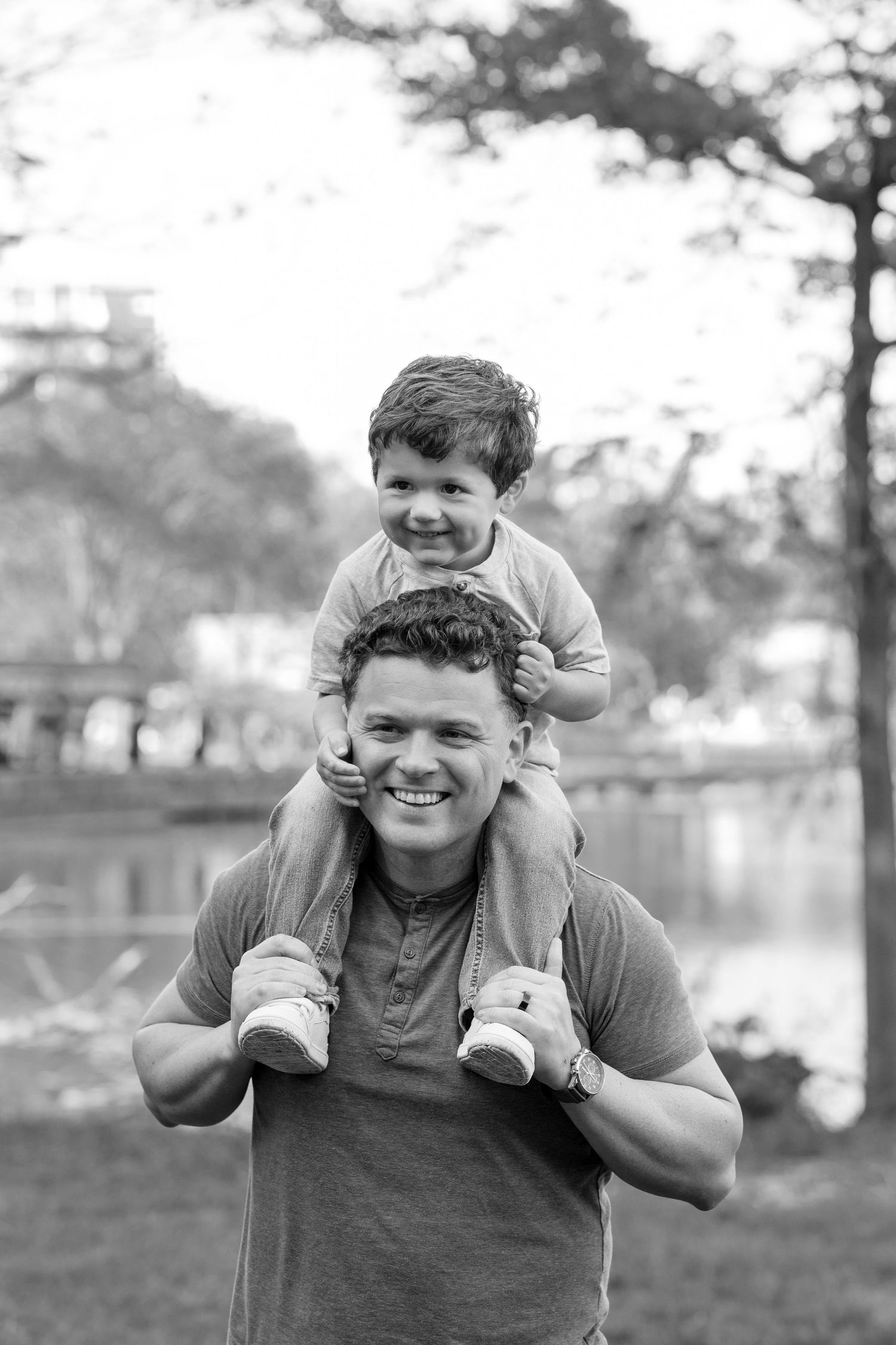 A smiling father carries his happy toddler son on his shoulders in a black and white park photo.