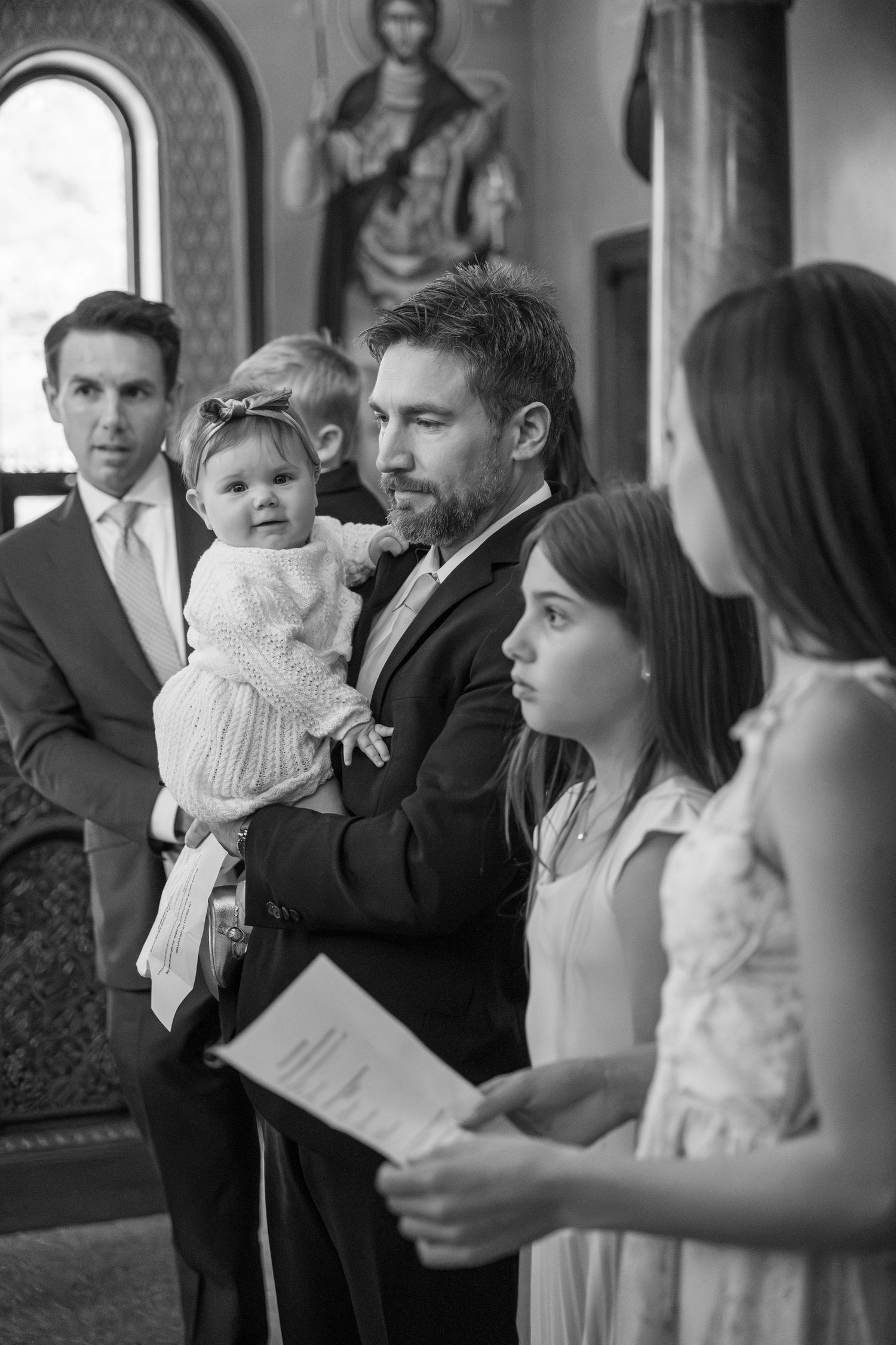 Black and white photo of a man holding a baby at a church baptism ceremony with family members nearby.