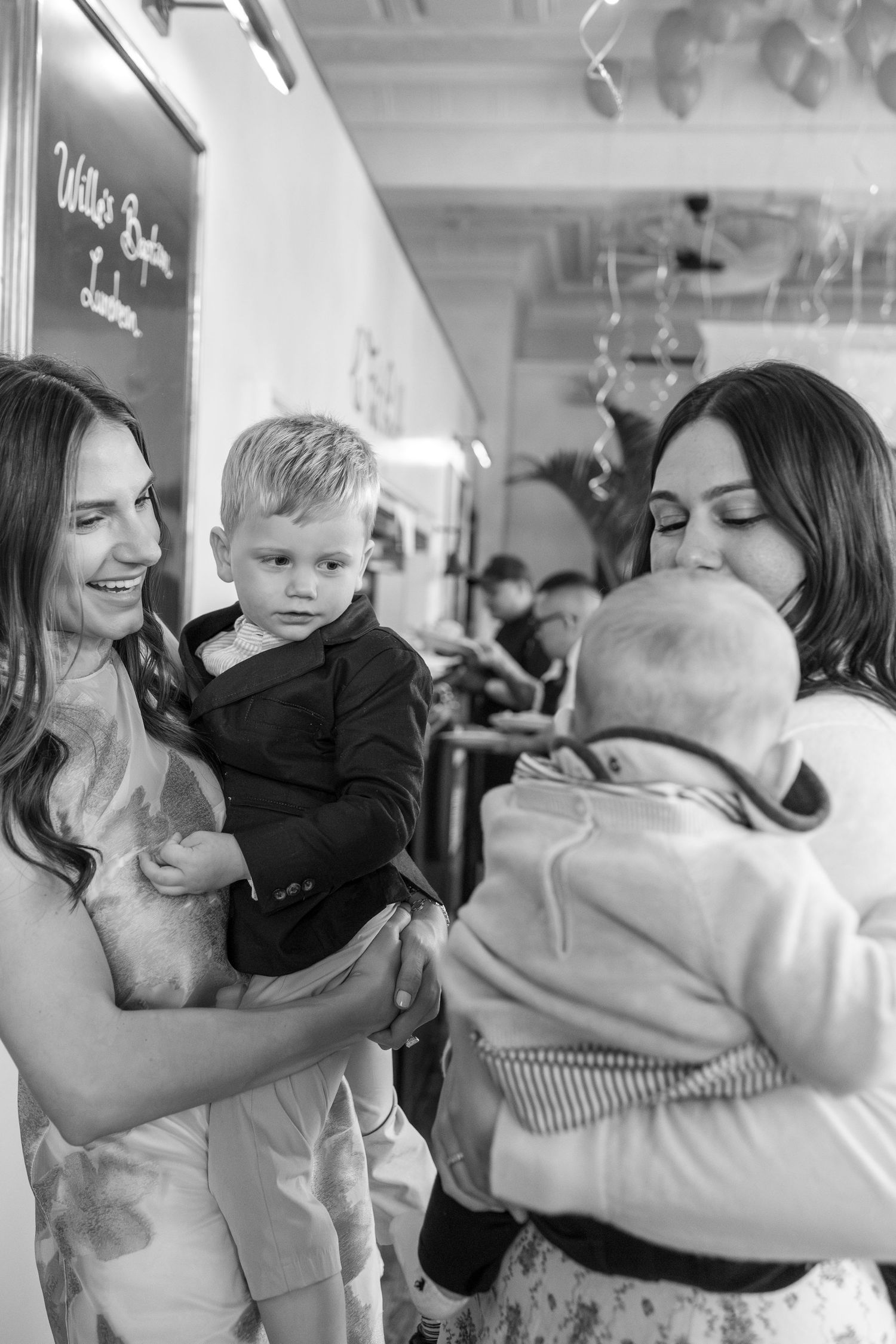 Two women holding young children at indoor celebration party in black and white photo.