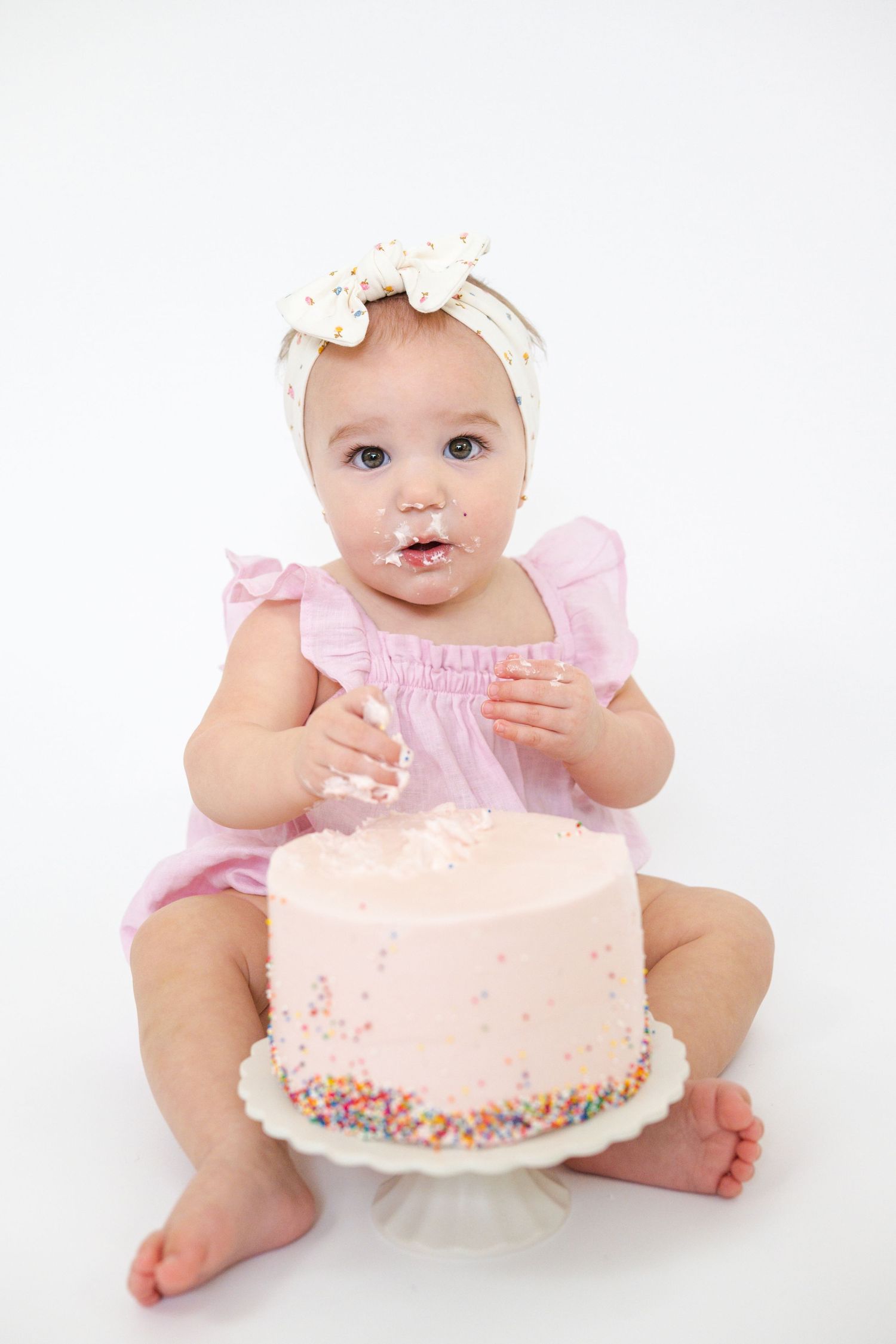 Baby in pink dress reaching for a light pink birthday cake with sprinkles.