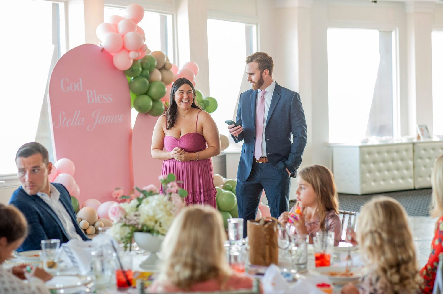 Wedding reception guests seated at tables in a bright room with pink decor and floral centerpieces.