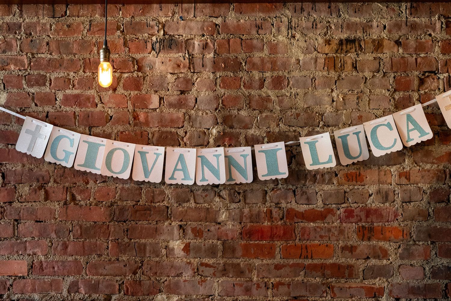 A white banner with the text 'Giovanni Luca' hangs against an exposed brick wall with pendant lighting.