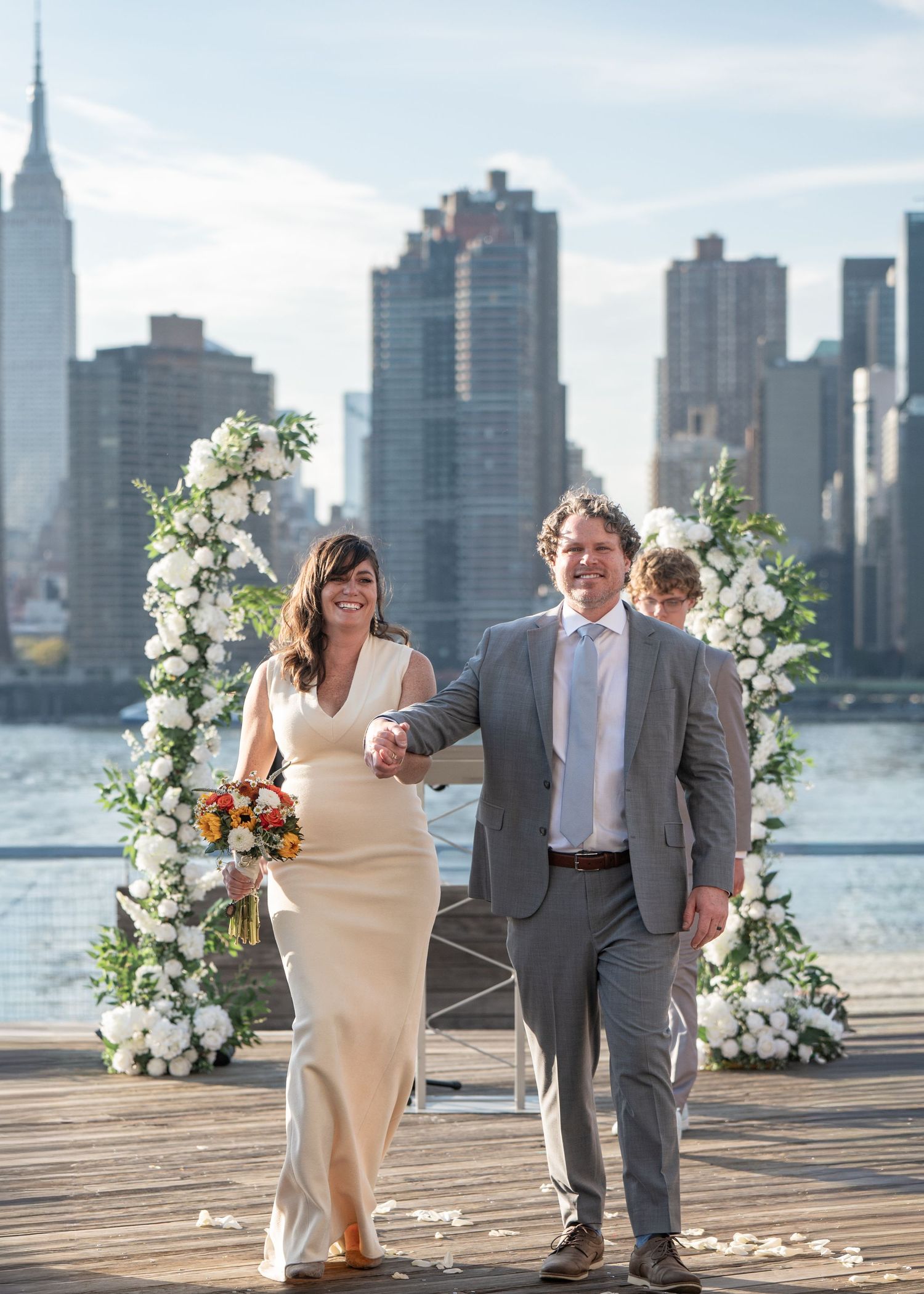A wedding couple poses on a waterfront deck with floral arrangements and the city skyline in the background.