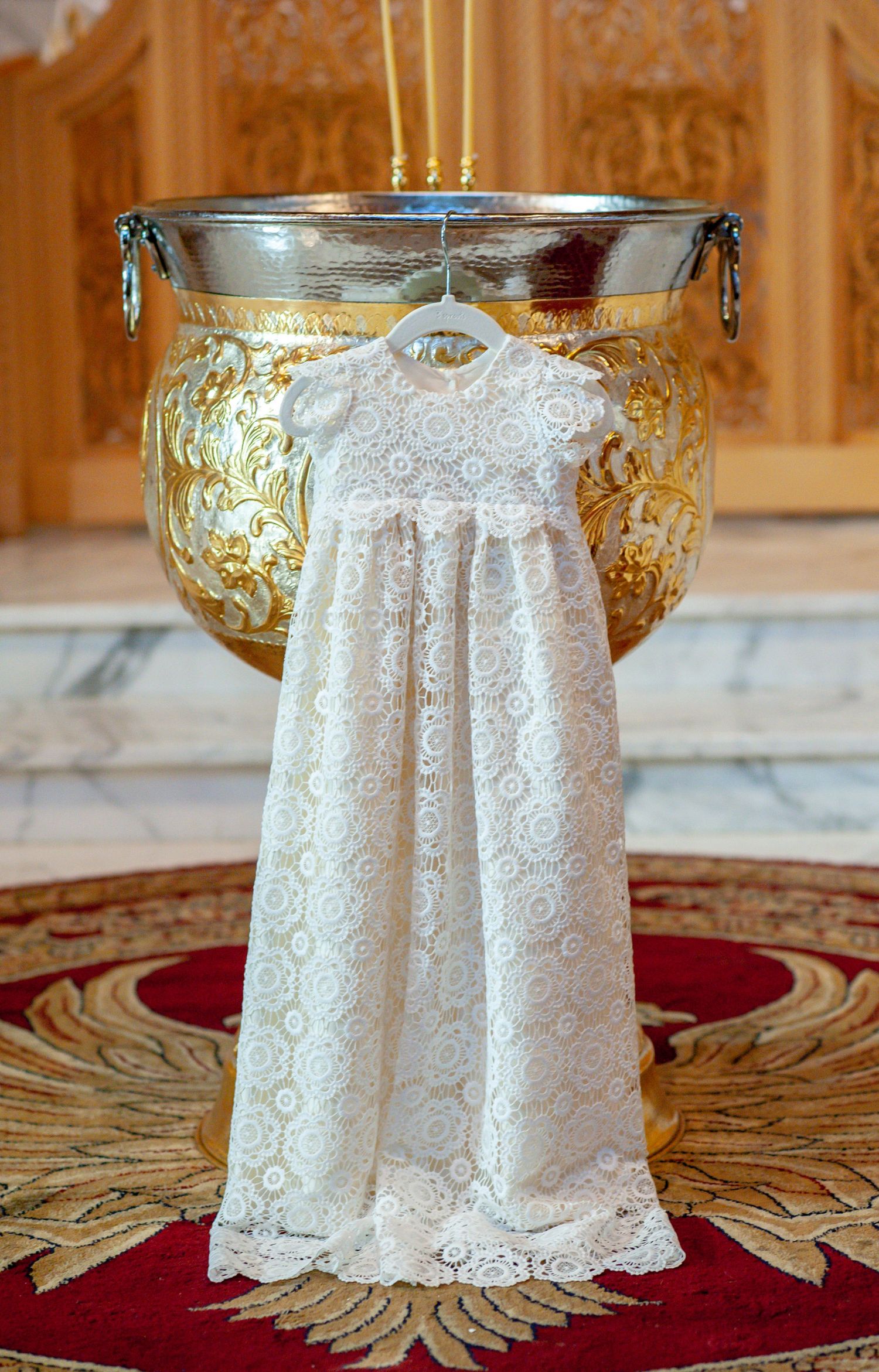 Delicate white lace christening gown displayed on a hanger over an ornate gold baptismal font in a church.