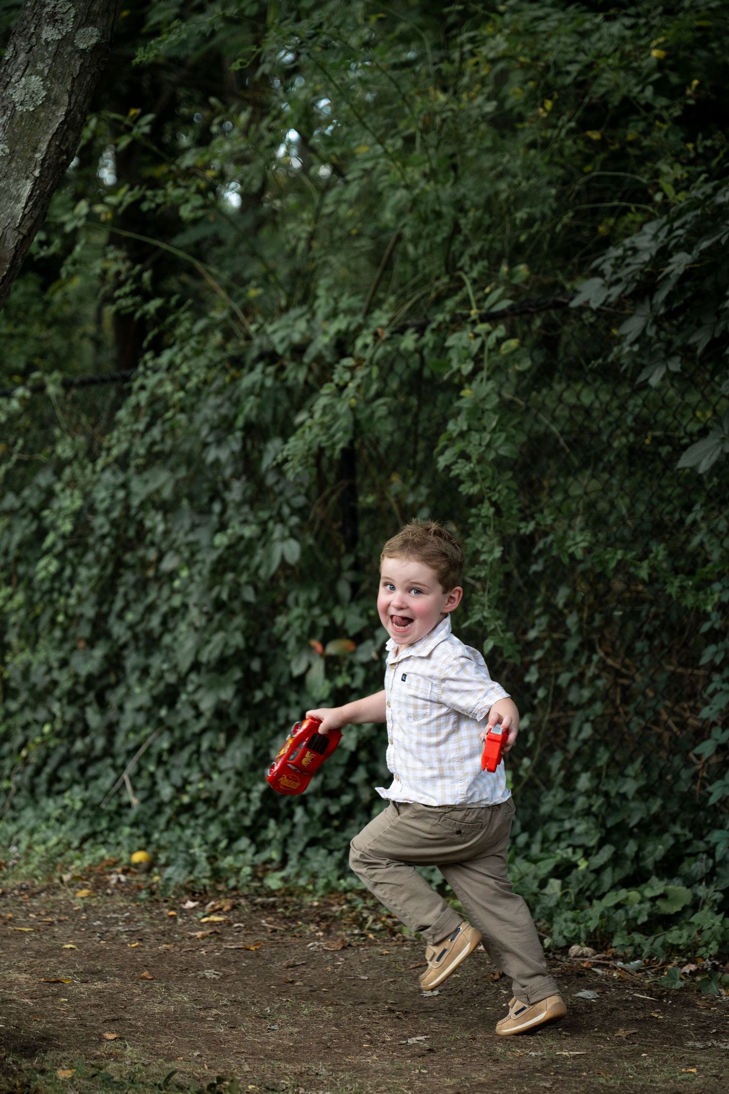 A child in khaki pants and striped shirt leaps playfully across a dirt path surrounded by green foliage.