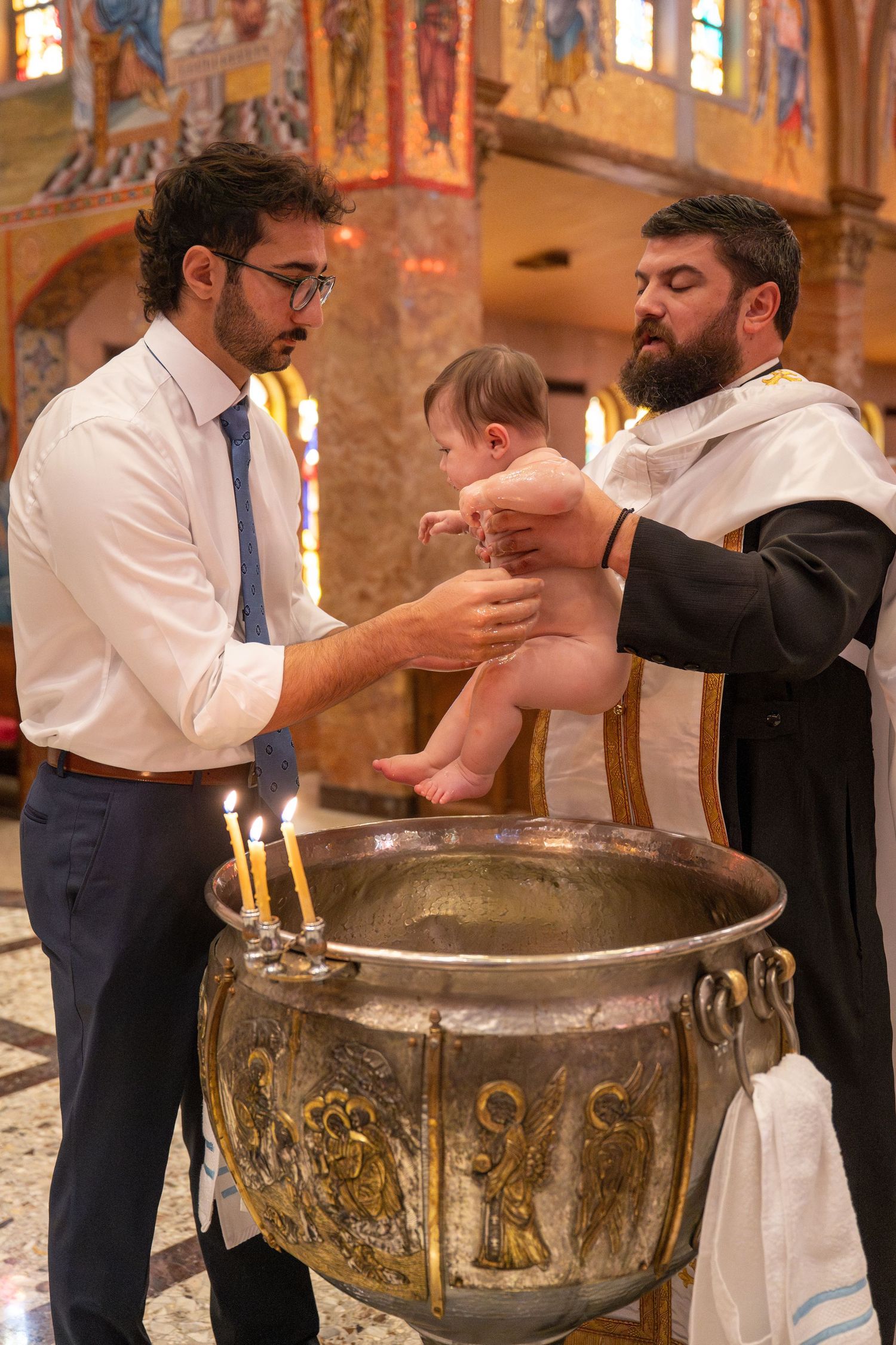 Priest performing baptism ritual at ornate metal baptismal font in church.
