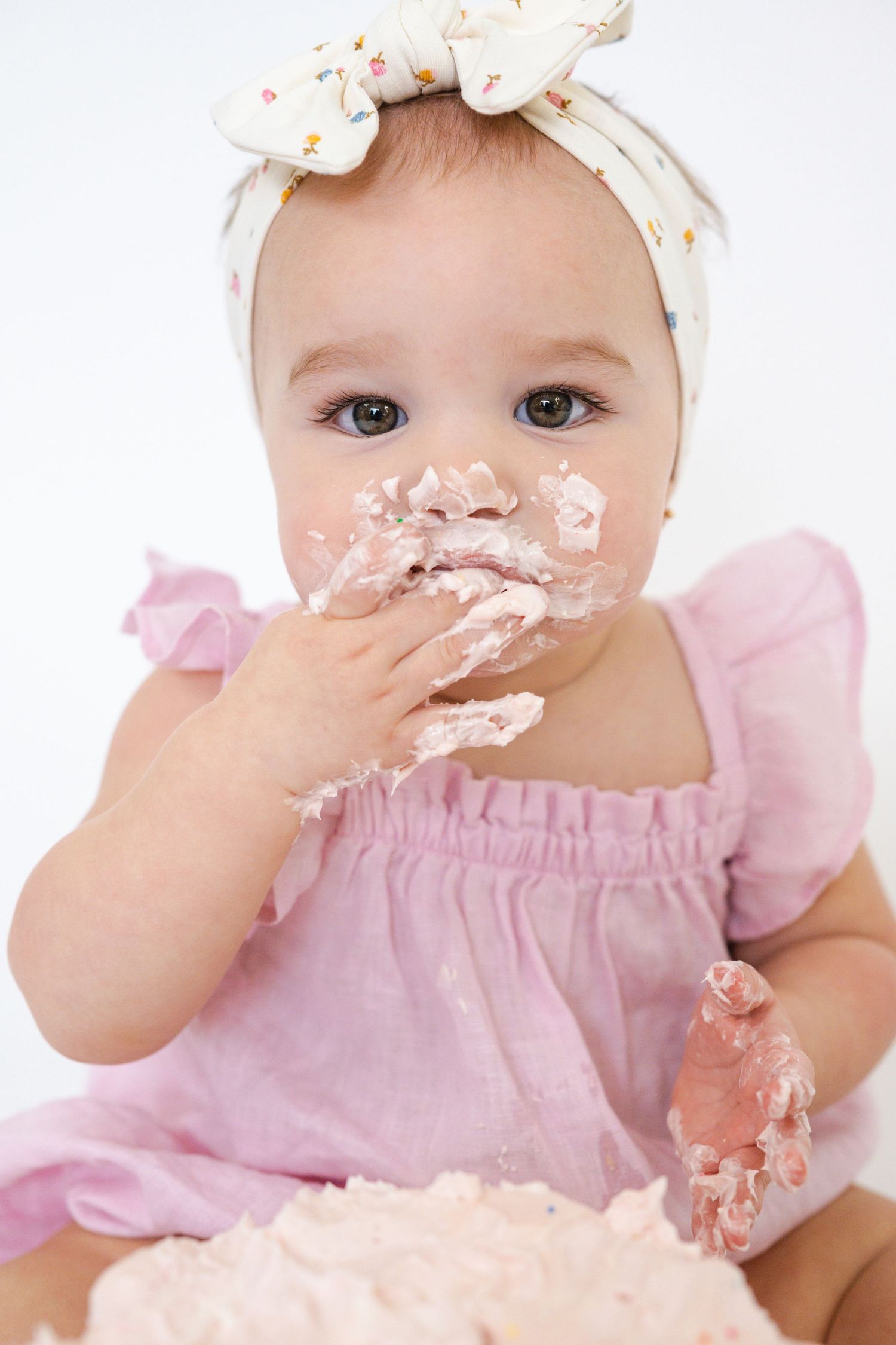 Baby in pink dress messily eating pink birthday cake during cake smash photoshoot.